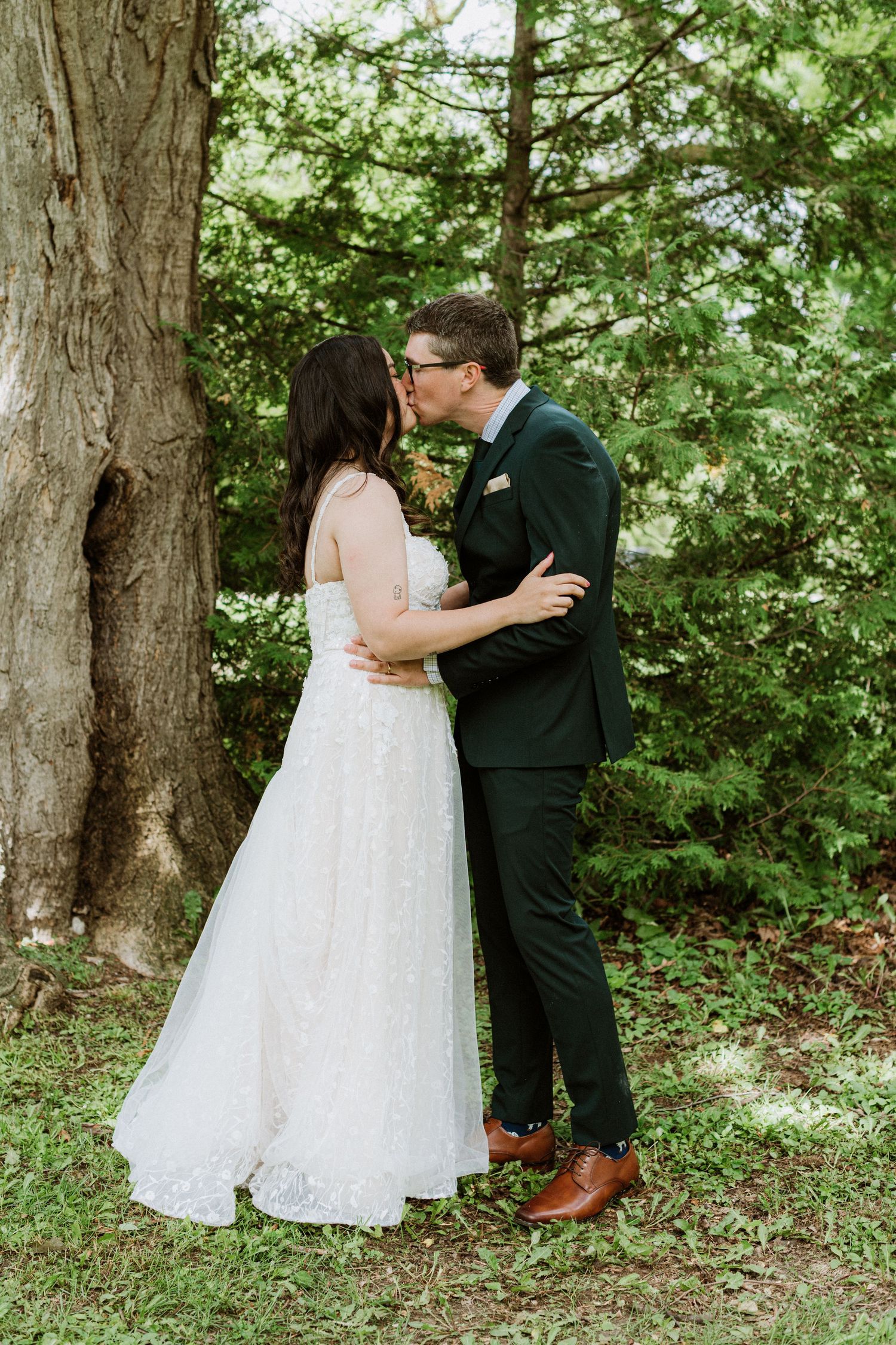 Bride and groom sharing romantic kiss beside large tree surrounded by lush green foliage.