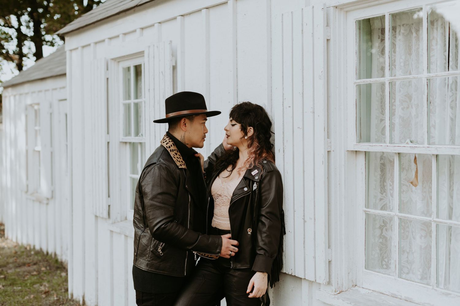 Couple in leather jackets sharing a tender gaze while leaning against a white wooden building.