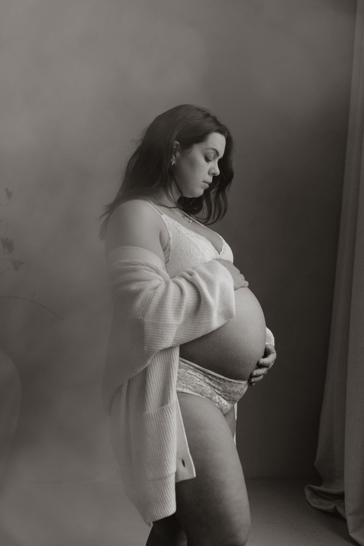 Black and white portrait of a pregnant woman in lace lingerie and cardigan cradling her bump.