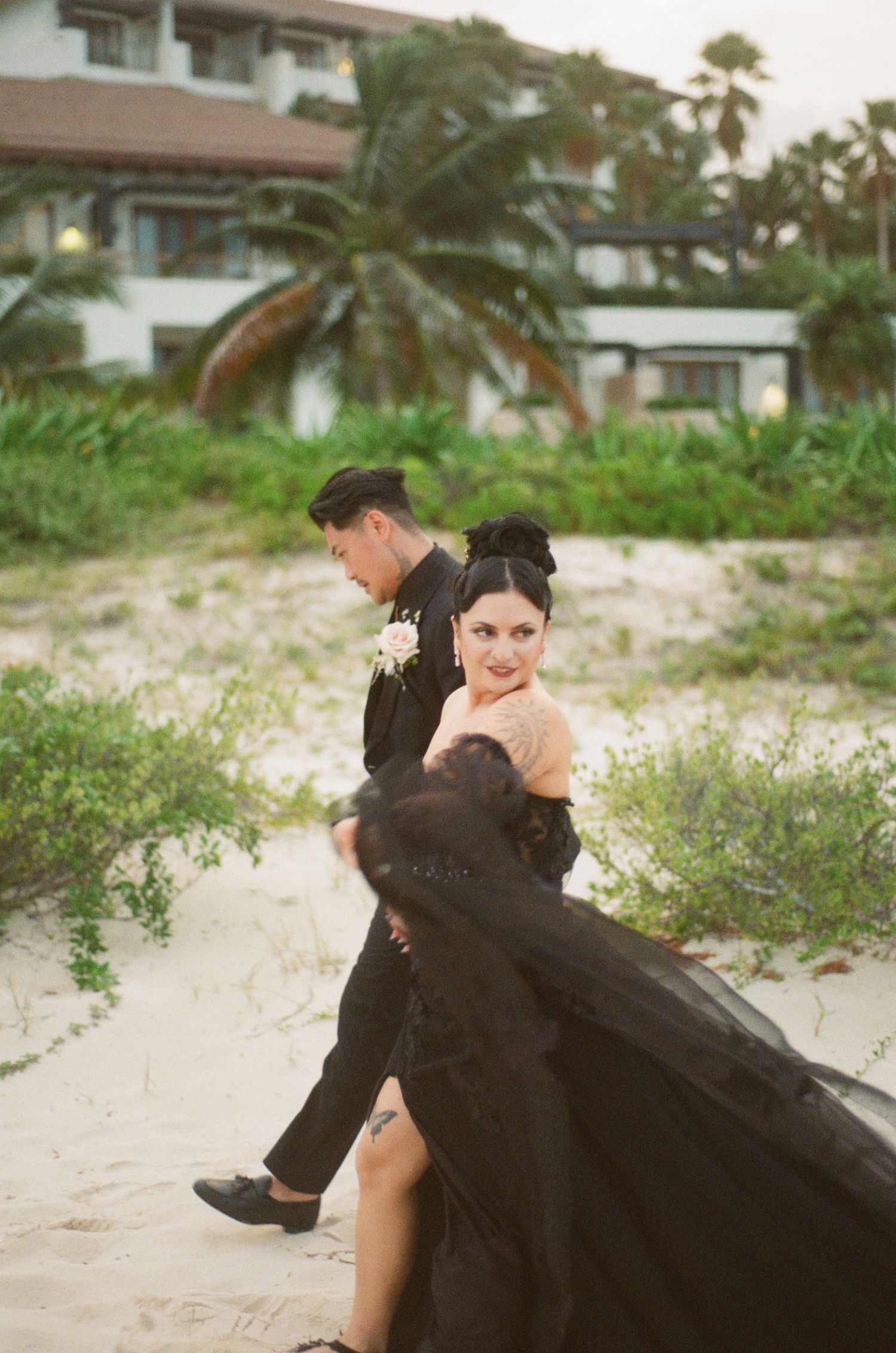 Bride in dramatic black gown looks back while groom leads her across sandy beach.