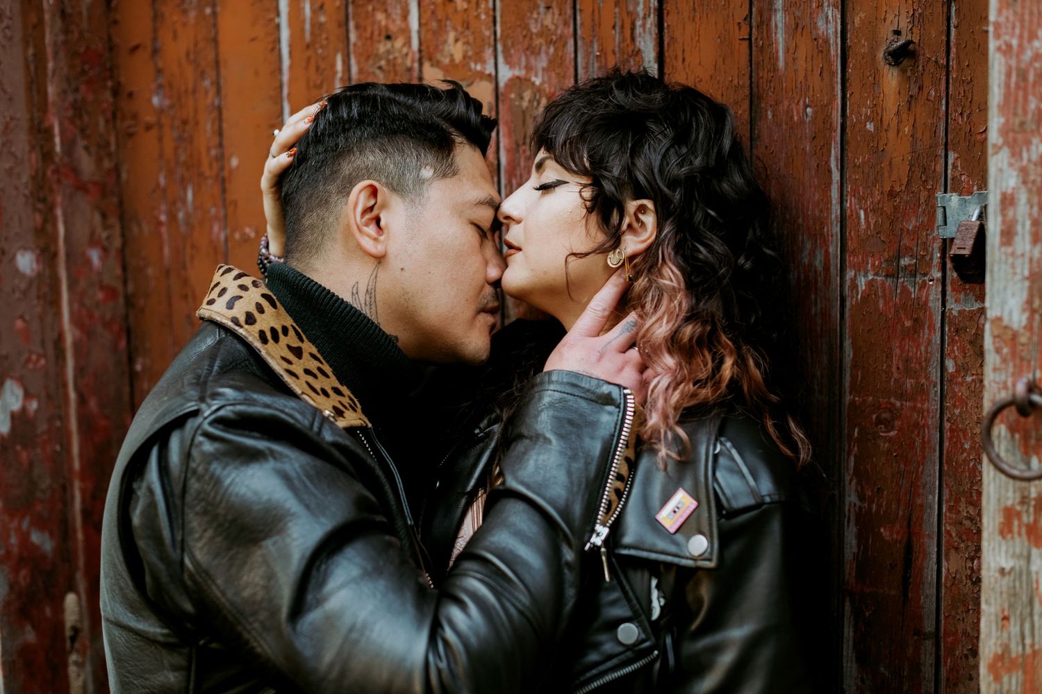 Couple in black leather jackets sharing an intimate kiss against a rustic wooden door.