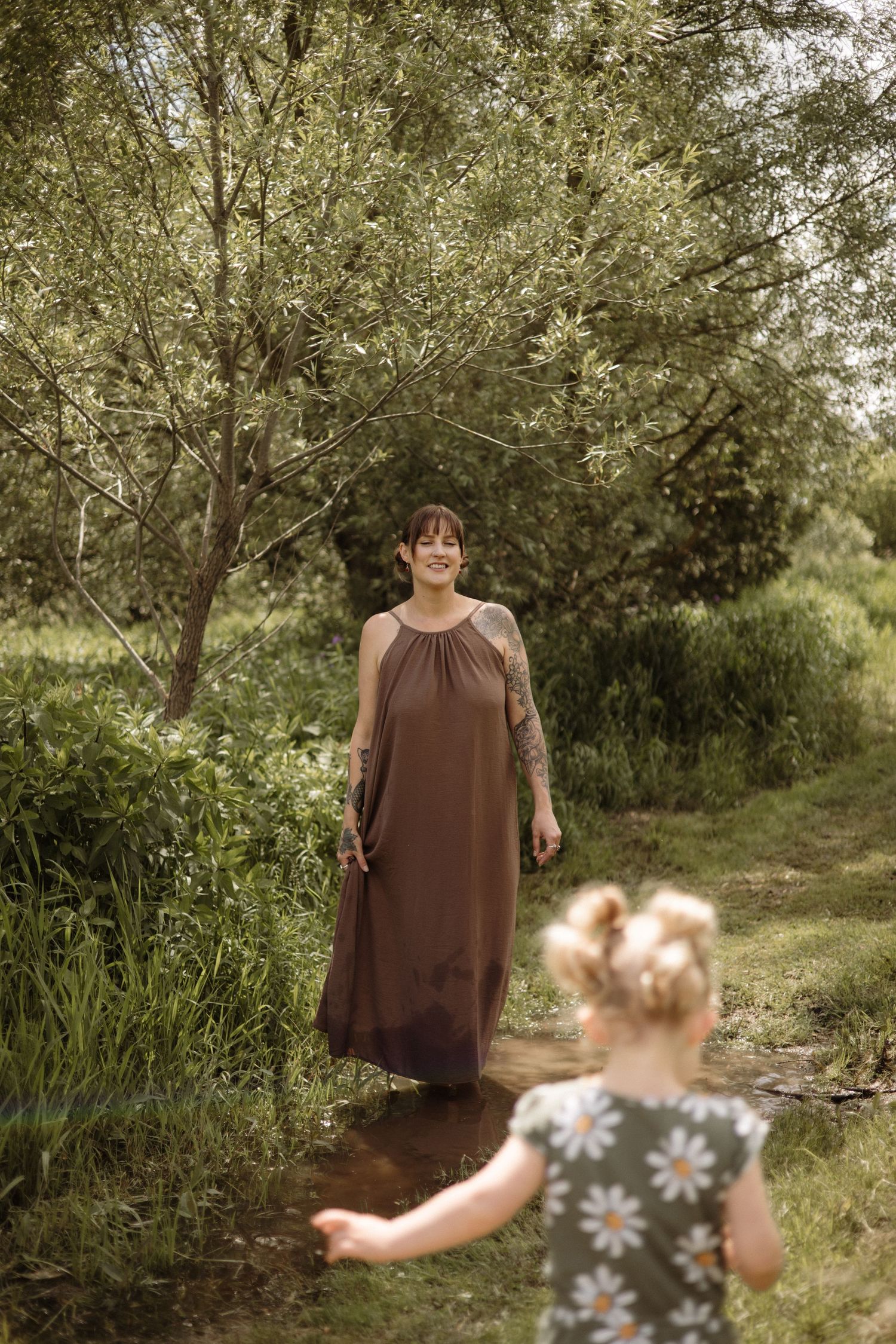 Smiling woman in brown dress stands in stream as toddler in daisy romper runs toward her.