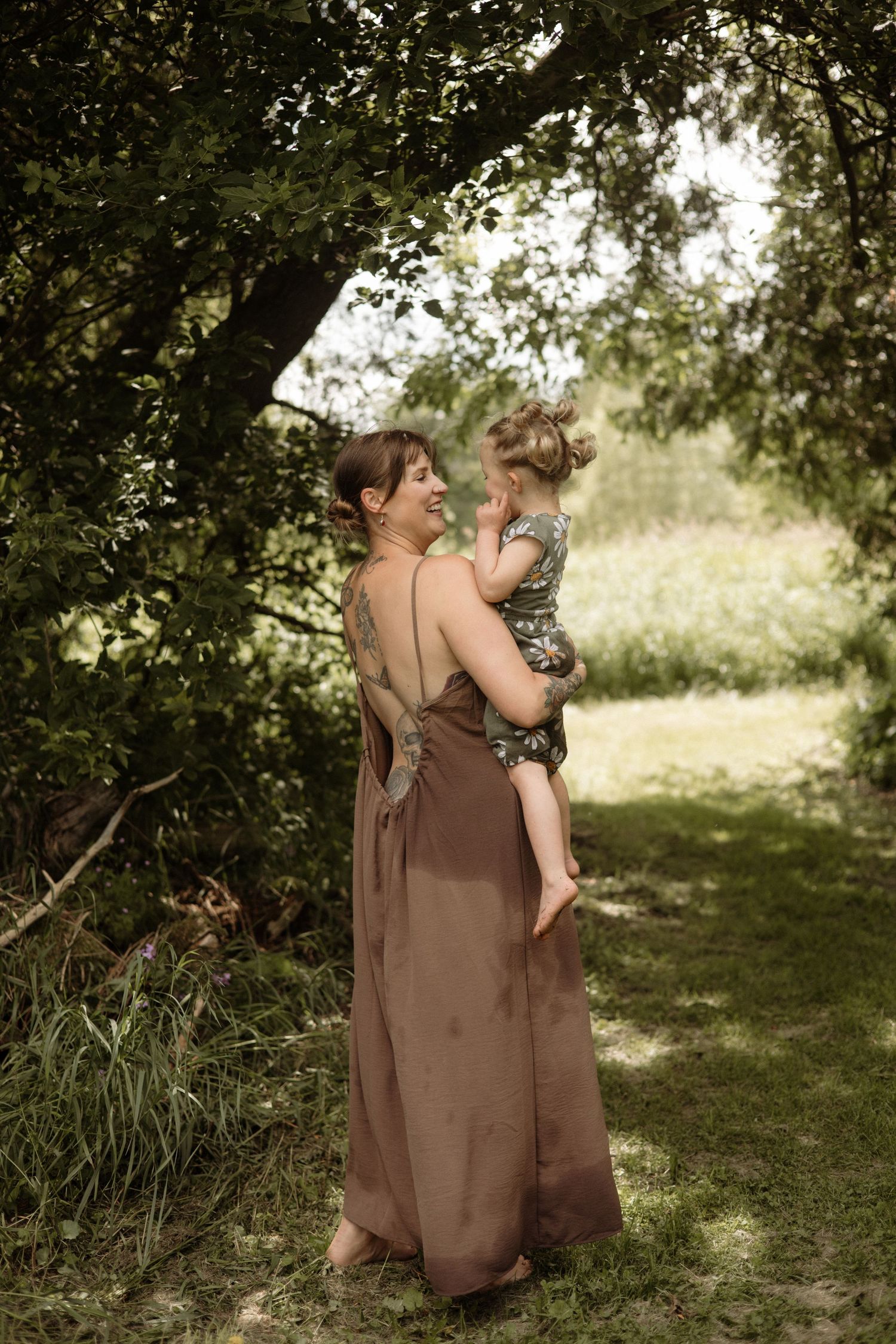 A smiling tattooed mother in a brown maxi dress holds her happy toddler daughter under green trees.