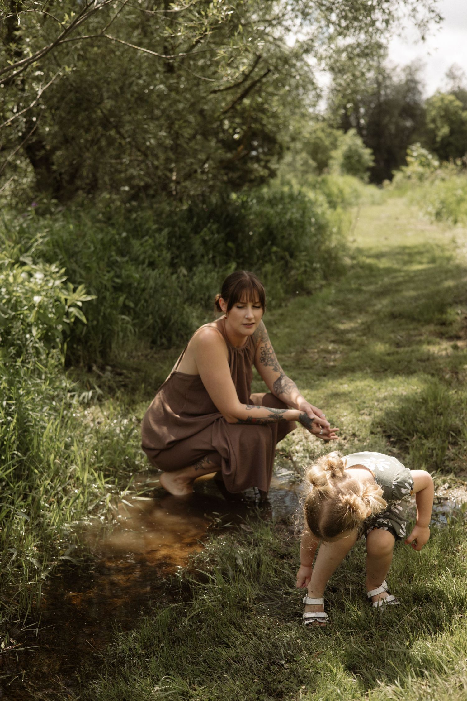 Tattooed mother in brown dress crouching by stream with toddler exploring water in lush green nature setting.