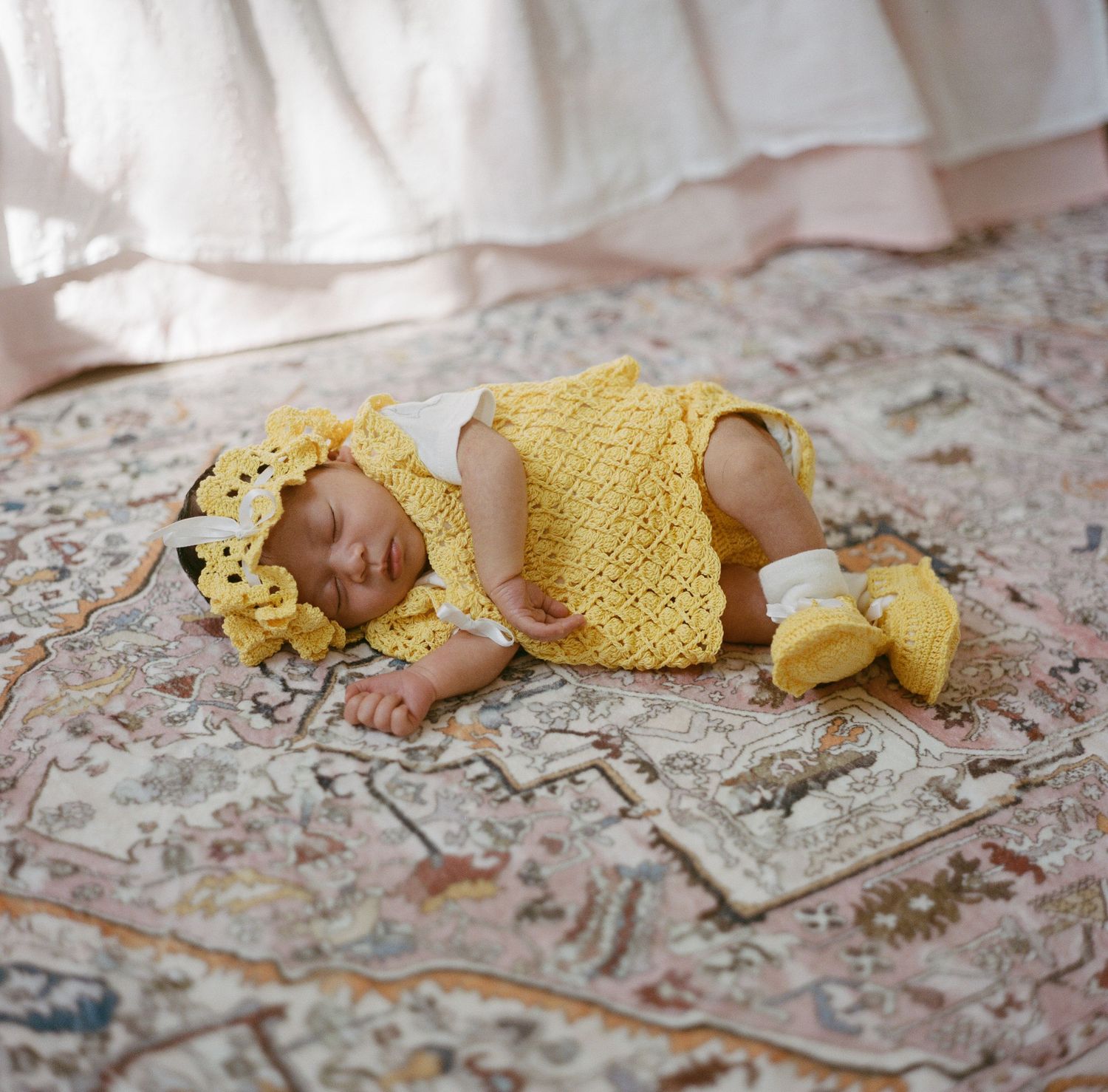 Sleeping newborn in yellow knit outfit and headband rests on a vintage floral rug.