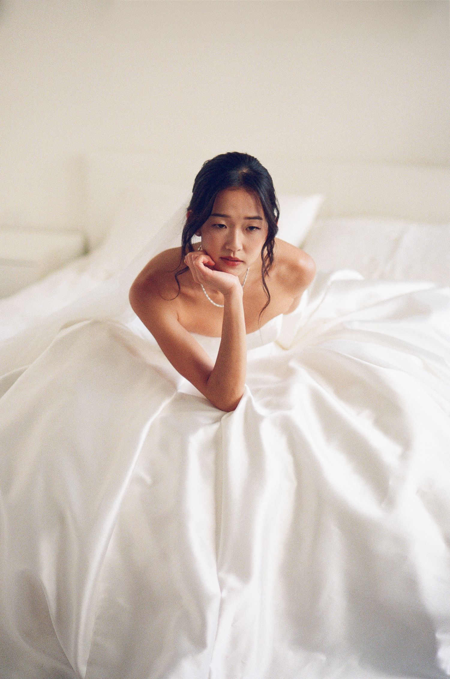 Asian bride in strapless white ball gown sitting on a bed, resting her chin on her hands.