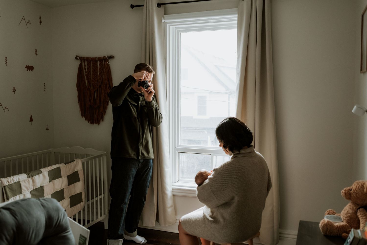 Father photographs mother holding newborn baby in cozy nursery room near window with natural light.