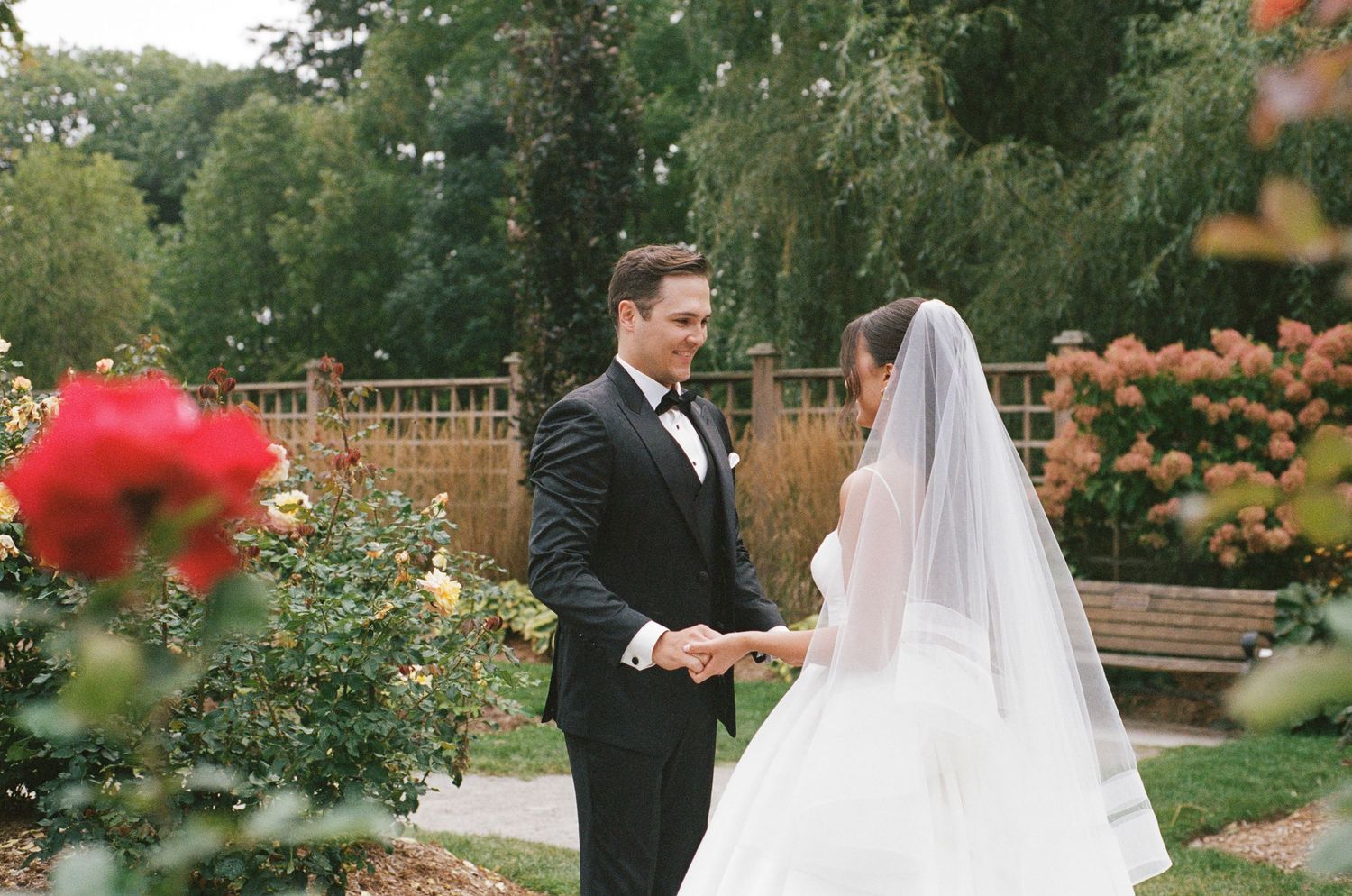 Groom in tuxedo and bride in white gown with veil holding hands during first look in rose garden.
