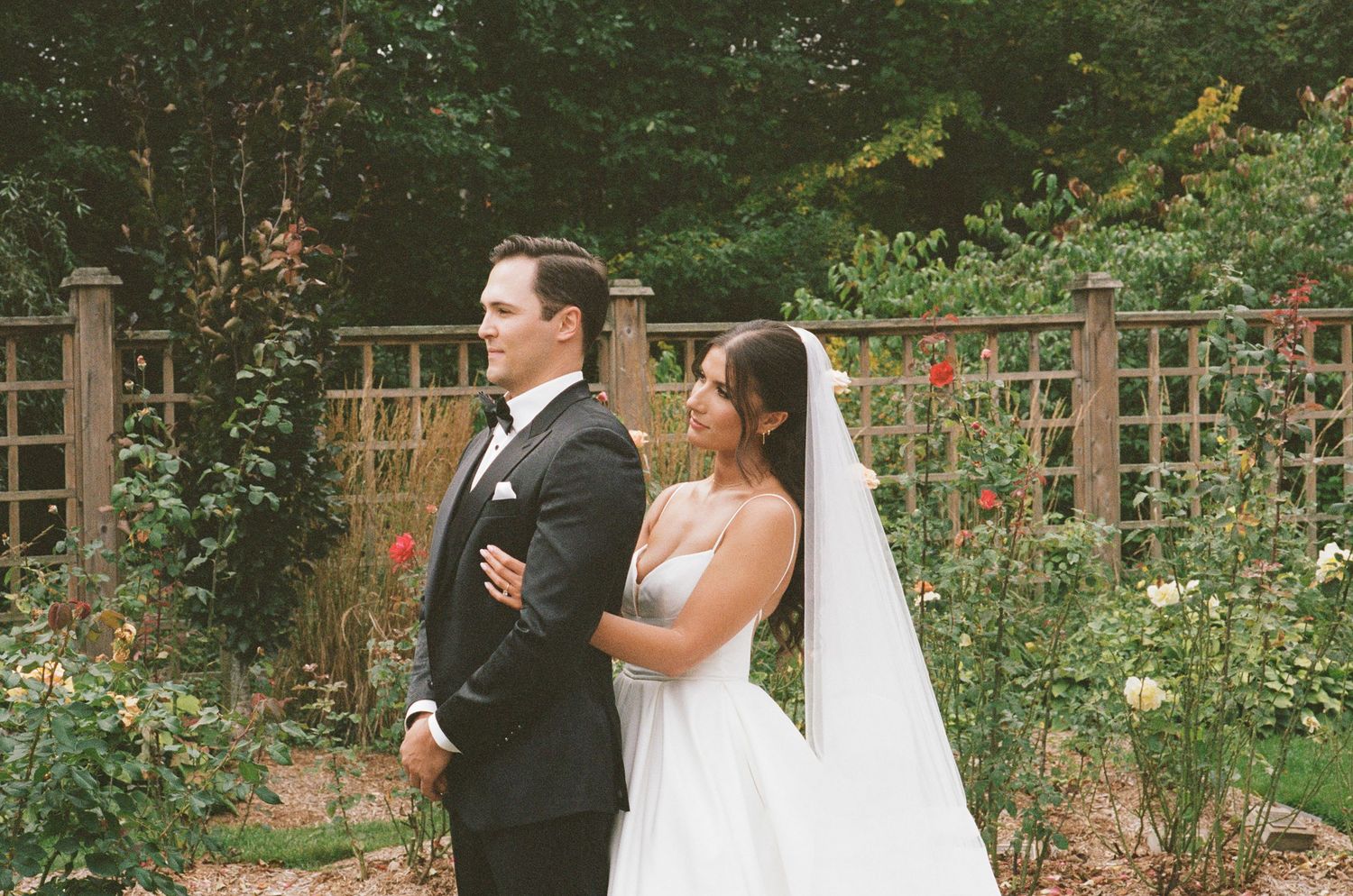 Bride and groom standing back to back in a rose garden with a wooden trellis fence backdrop.