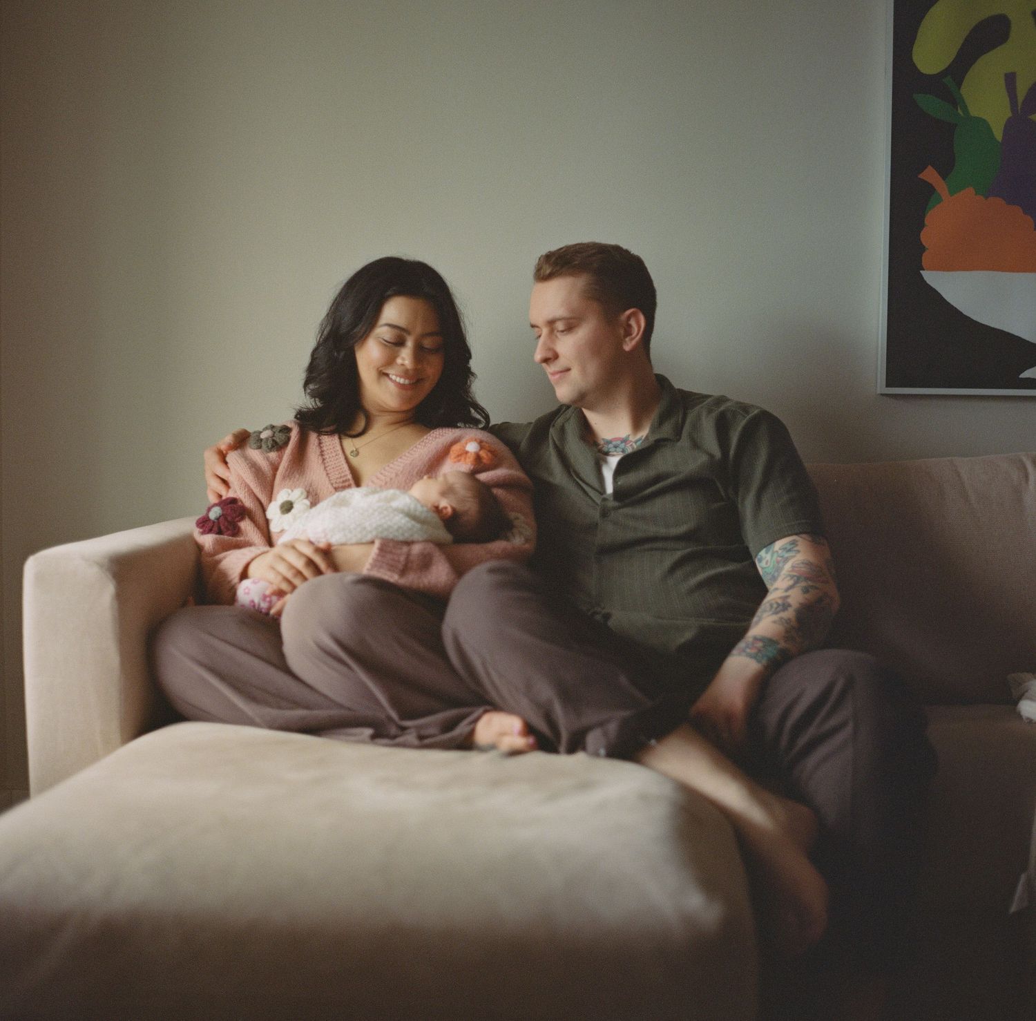 Happy couple with tattoos sitting on beige sofa holding their sleeping newborn baby at home.