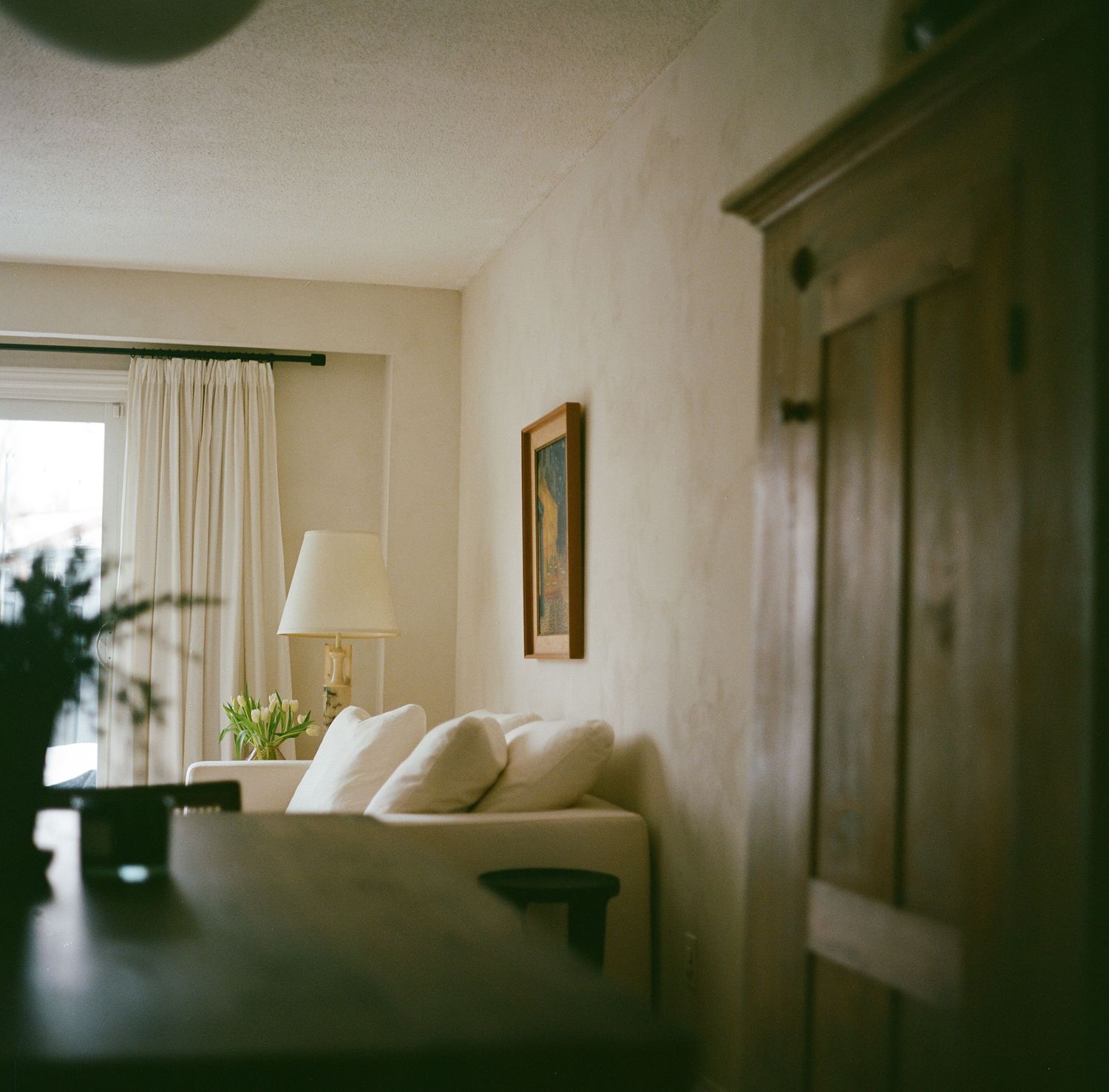 Cozy neutral living room viewed through doorway with white sofa, lamp, tulips, and framed artwork on wall.