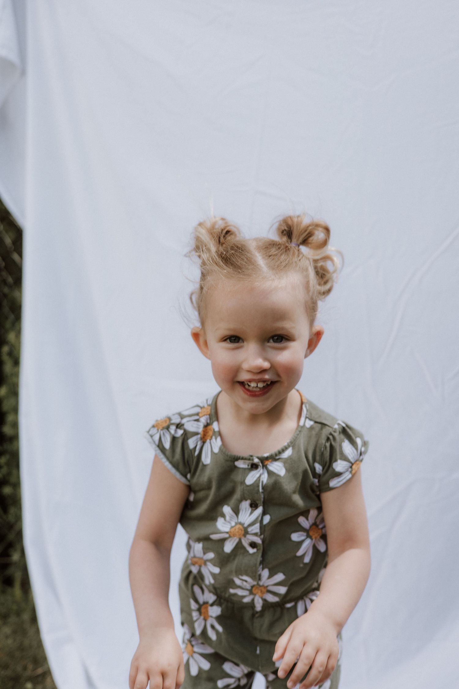 Happy toddler girl with pigtail buns wearing olive green daisy print romper smiling in front of white backdrop.