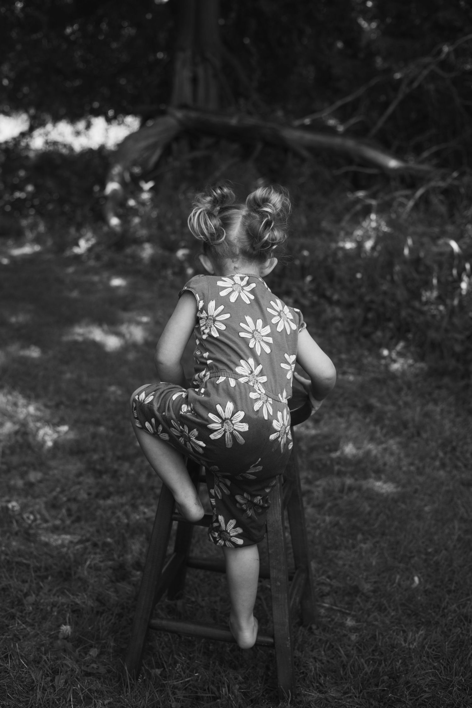 Black and white rear view of toddler girl in floral romper sitting on wooden stool in outdoor garden setting.