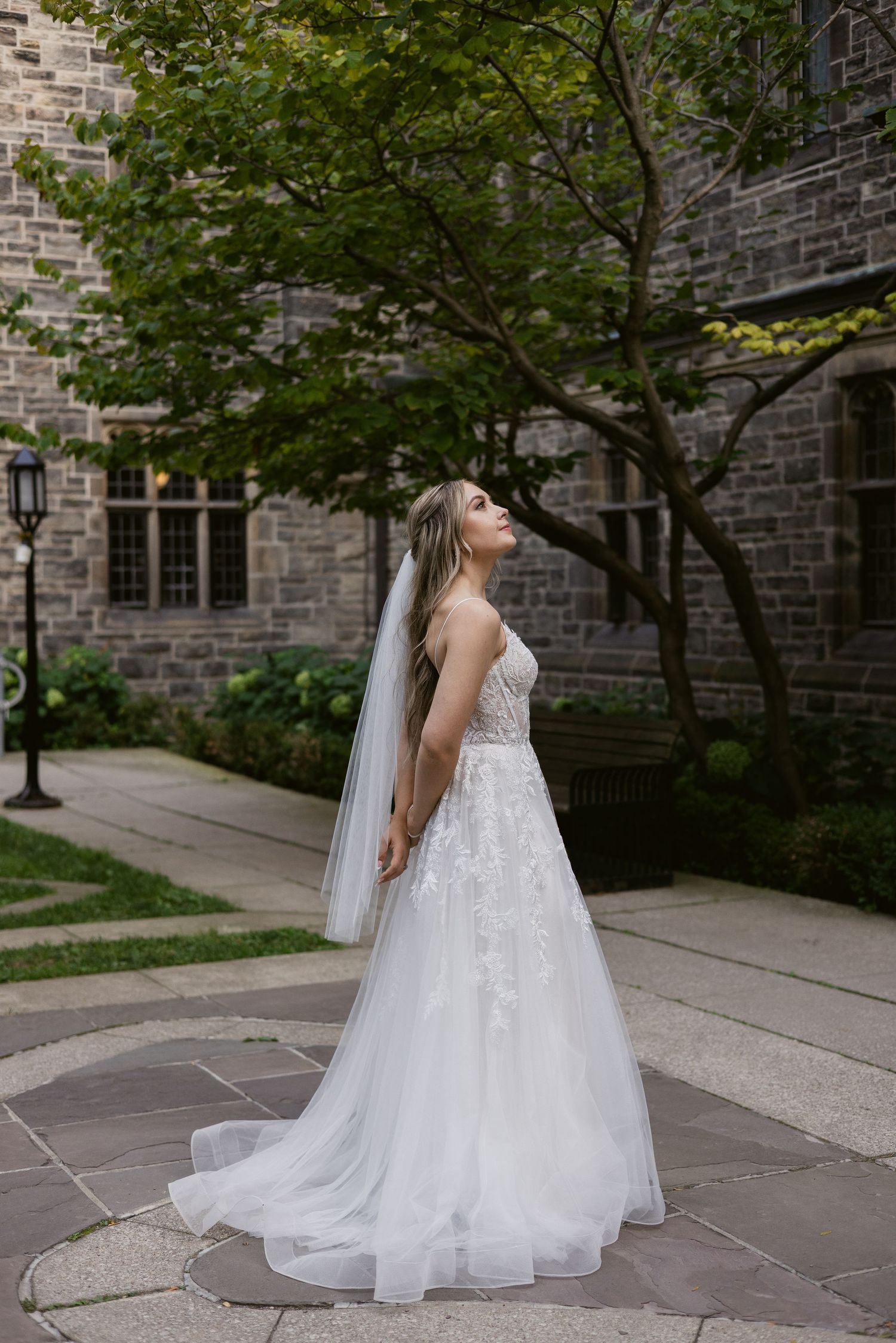 Bride in strapless lace gown and veil gazes upward beside a lush tree against a stone building backdrop.