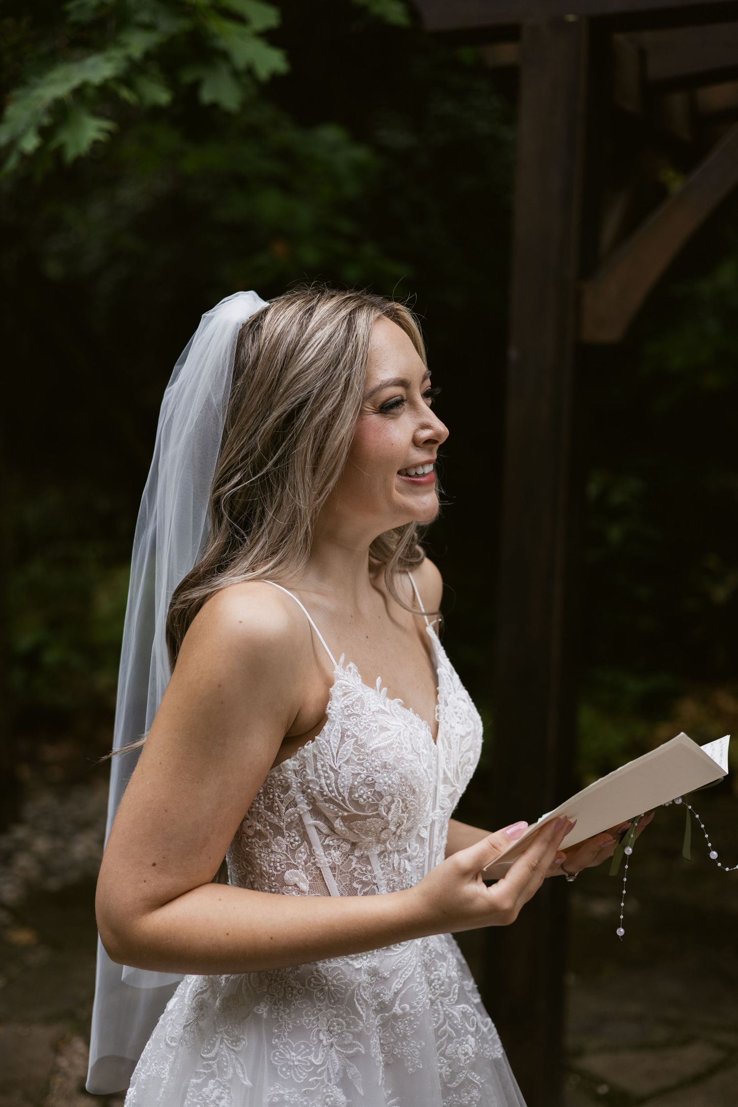 Joyful bride in lace wedding dress and veil laughing while reading vows at outdoor ceremony.