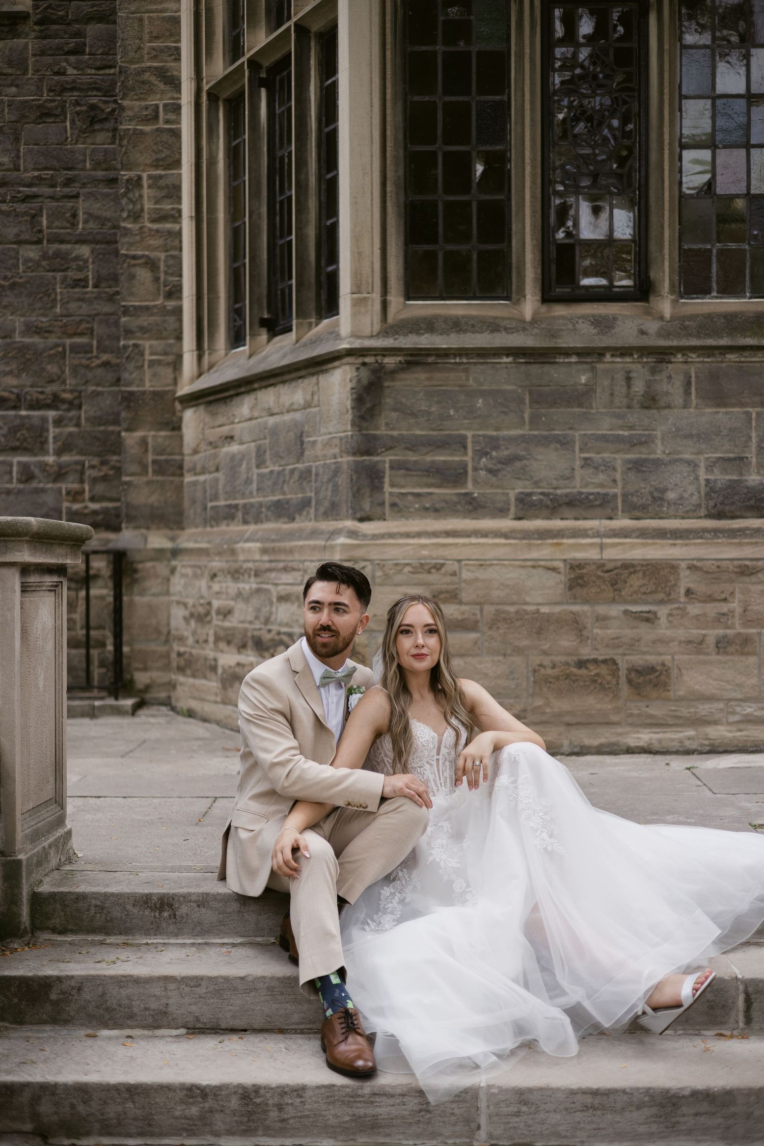 Bride and groom sit closely together on stone steps outside a grand historic stone building.