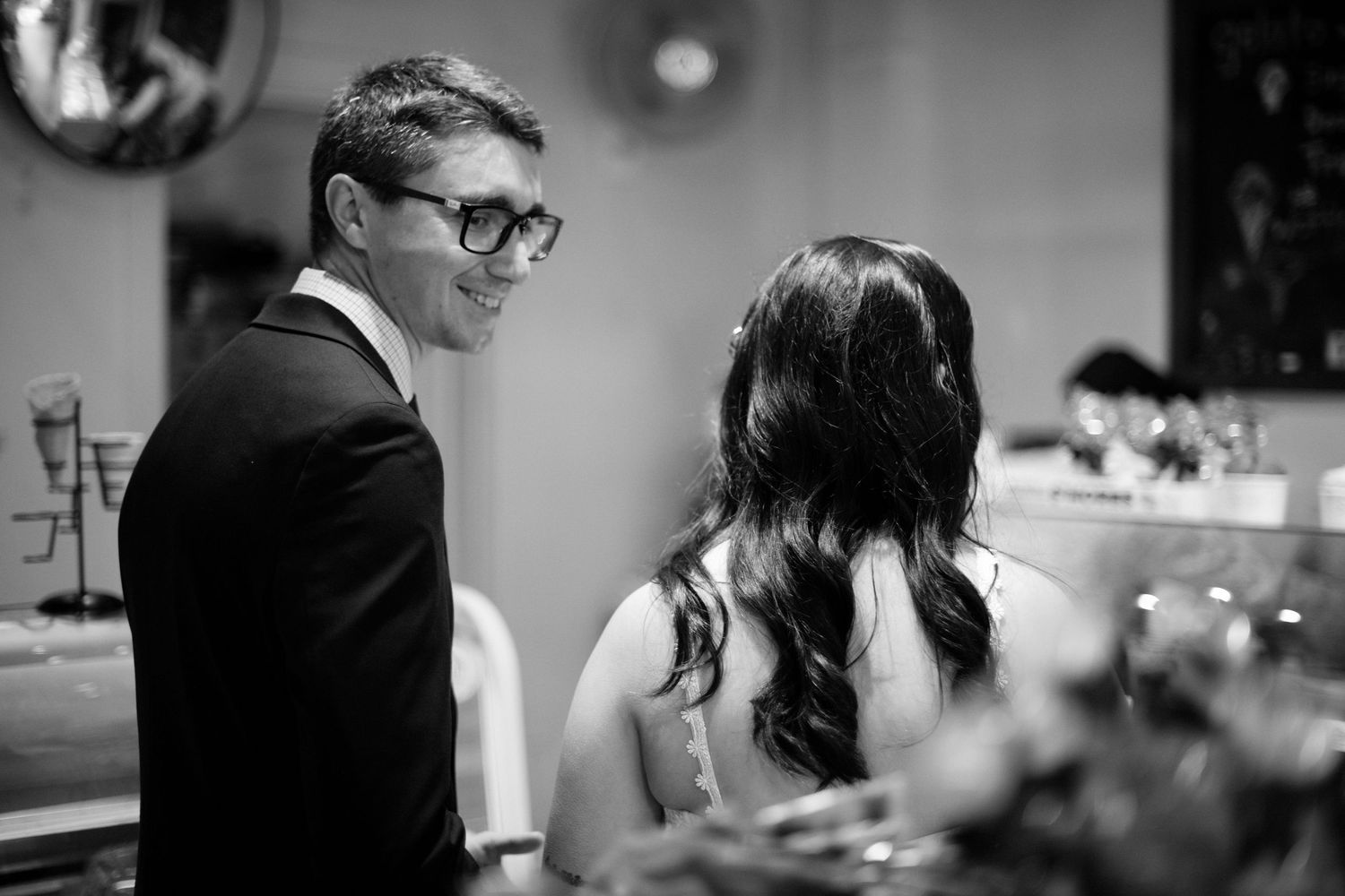 Black and white photo of groom in glasses smiling at bride during wedding reception dinner.