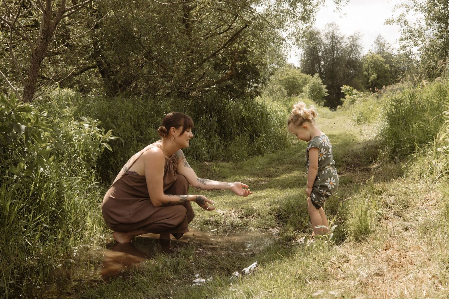 A mother and toddler play together beside a small stream in a sunny green outdoor setting.