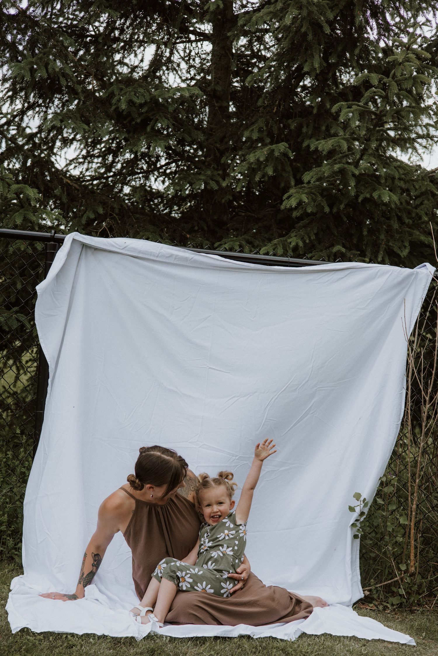 A mother and her toddler daughter sit together in front of a white backdrop in an outdoor garden.