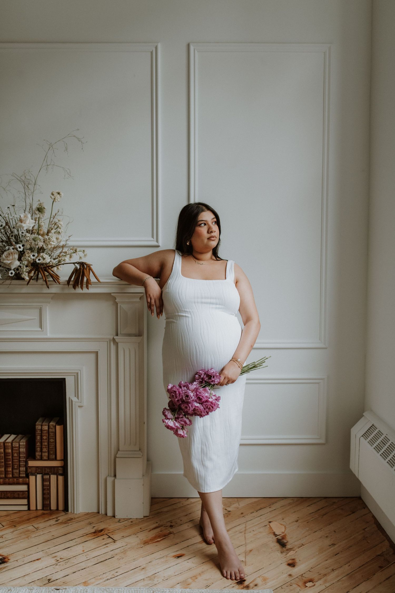 Pregnant woman in white ribbed dress holding pink tulips, leaning against fireplace mantel in elegant white room.