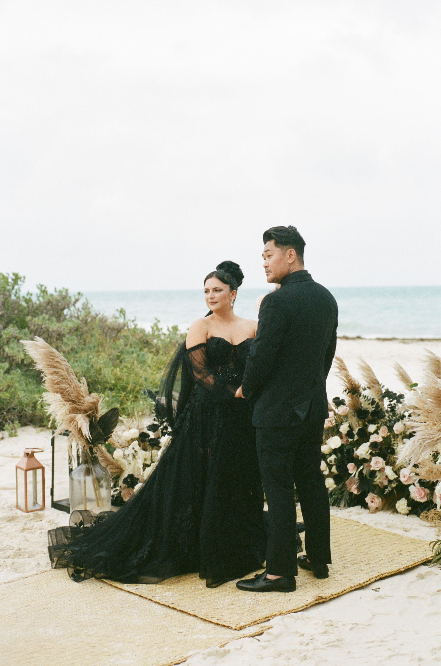 Bride and groom in matching black wedding attire standing at a beach altar with pampas grass and floral arrangements.