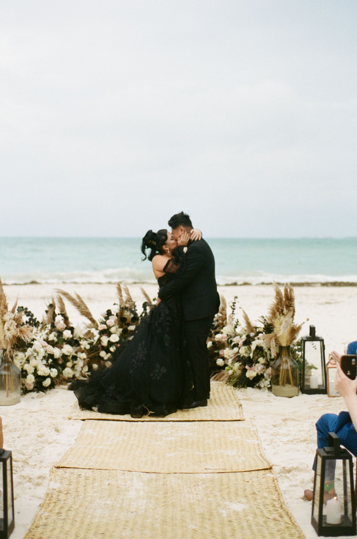 Newlyweds sharing first kiss at beach altar with white floral arrangements, pampas grass, and lanterns.
