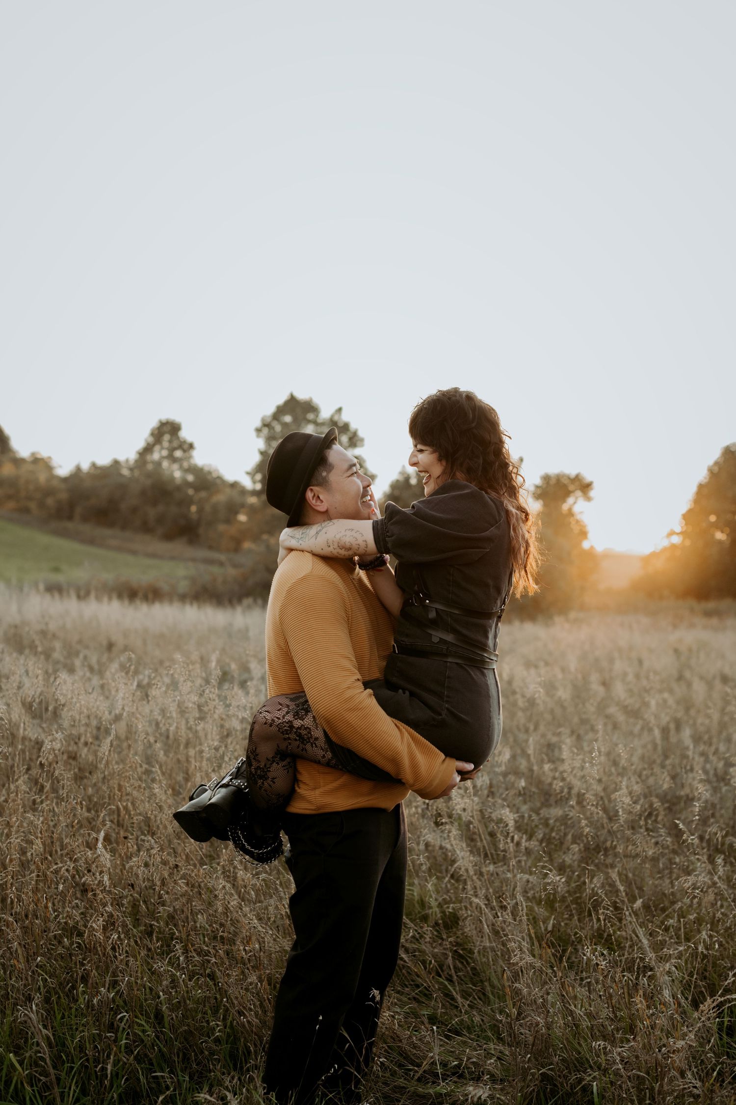 Man in mustard sweater lifting smiling woman in dark outfit in a glowing sunset meadow field.