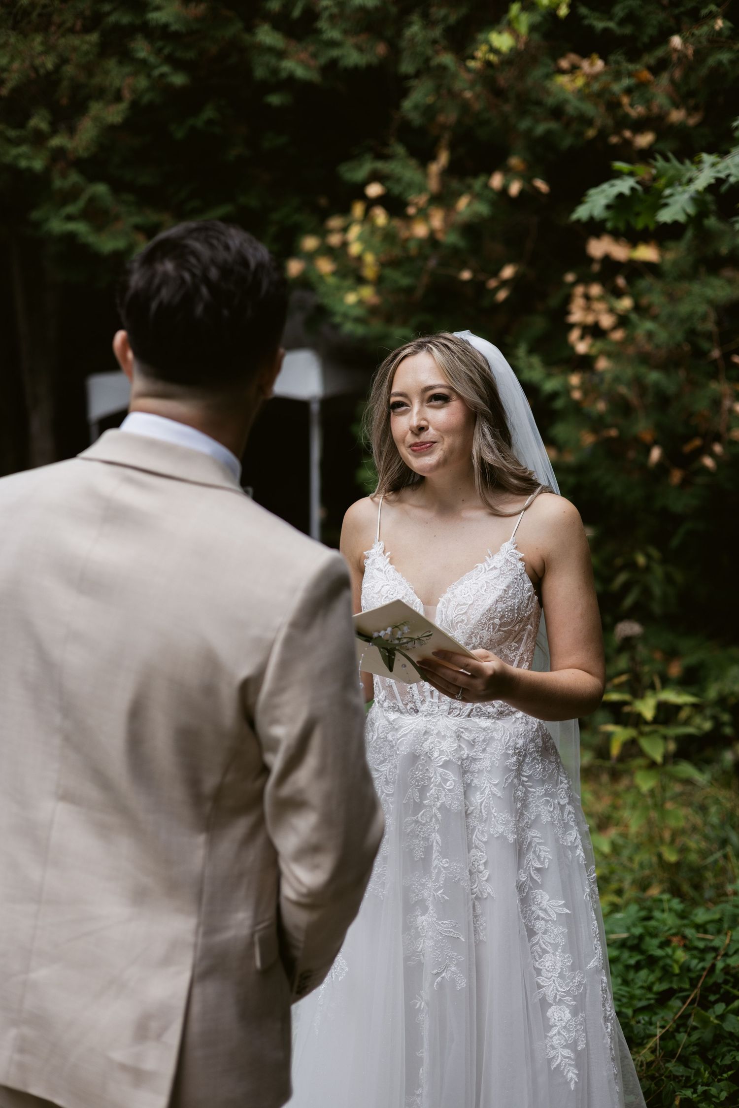 Radiant bride in lace gown holding vow booklet smiling at groom during outdoor wedding ceremony.