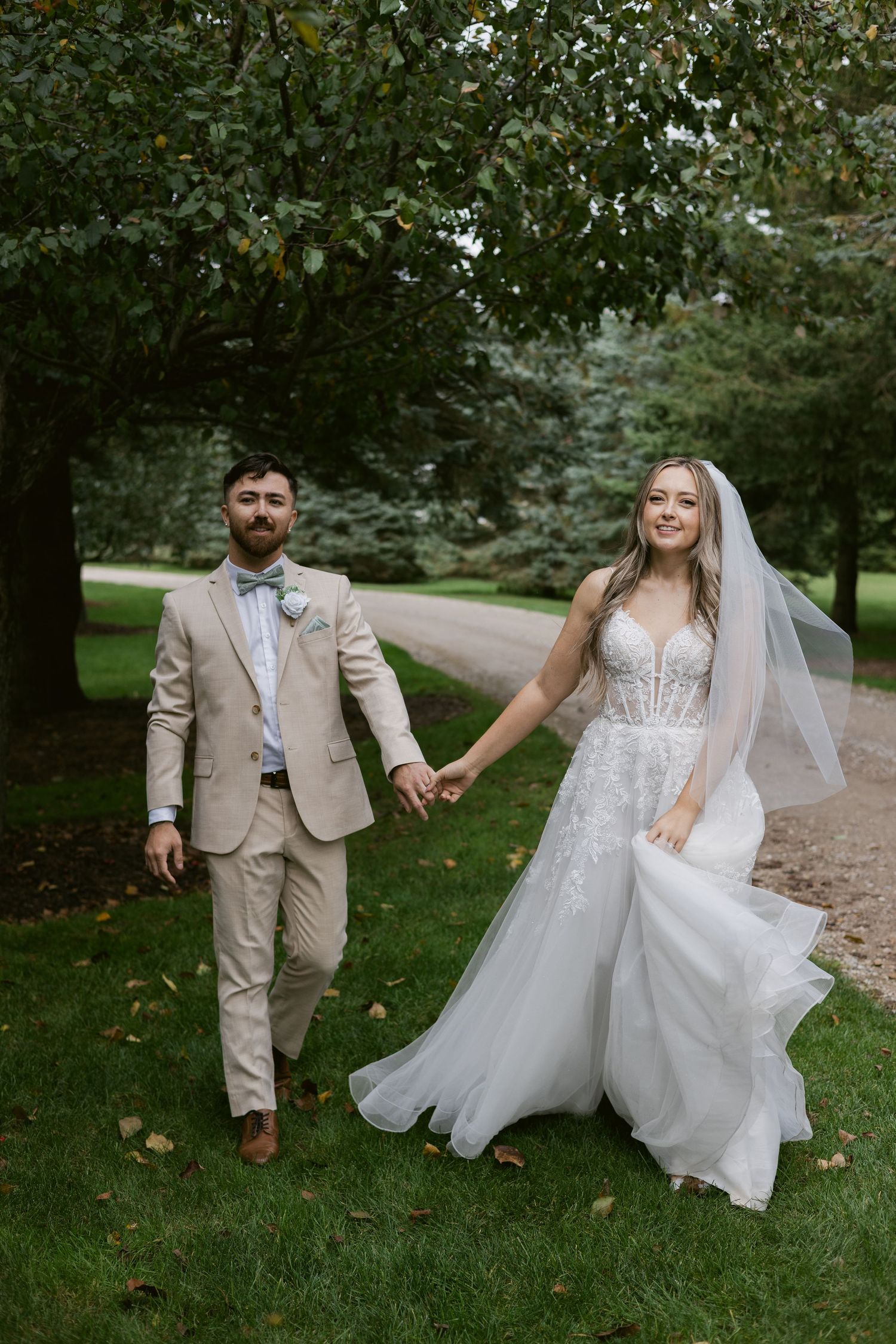 Smiling bride in lace gown and veil walks hand in hand with groom in beige suit on garden path.