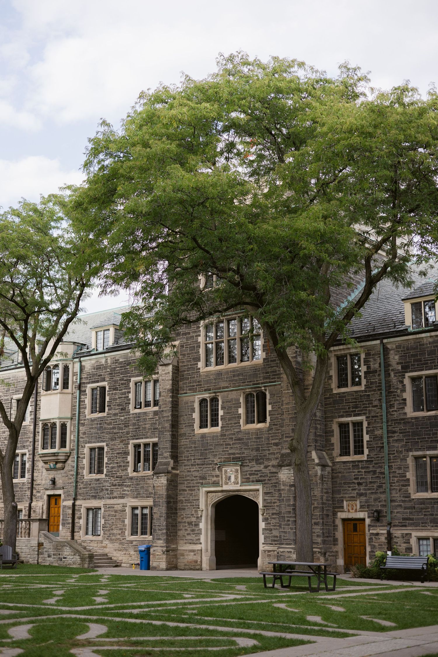 Gothic stone university building with arched entrance framed by large mature trees and decorative courtyard lawn.