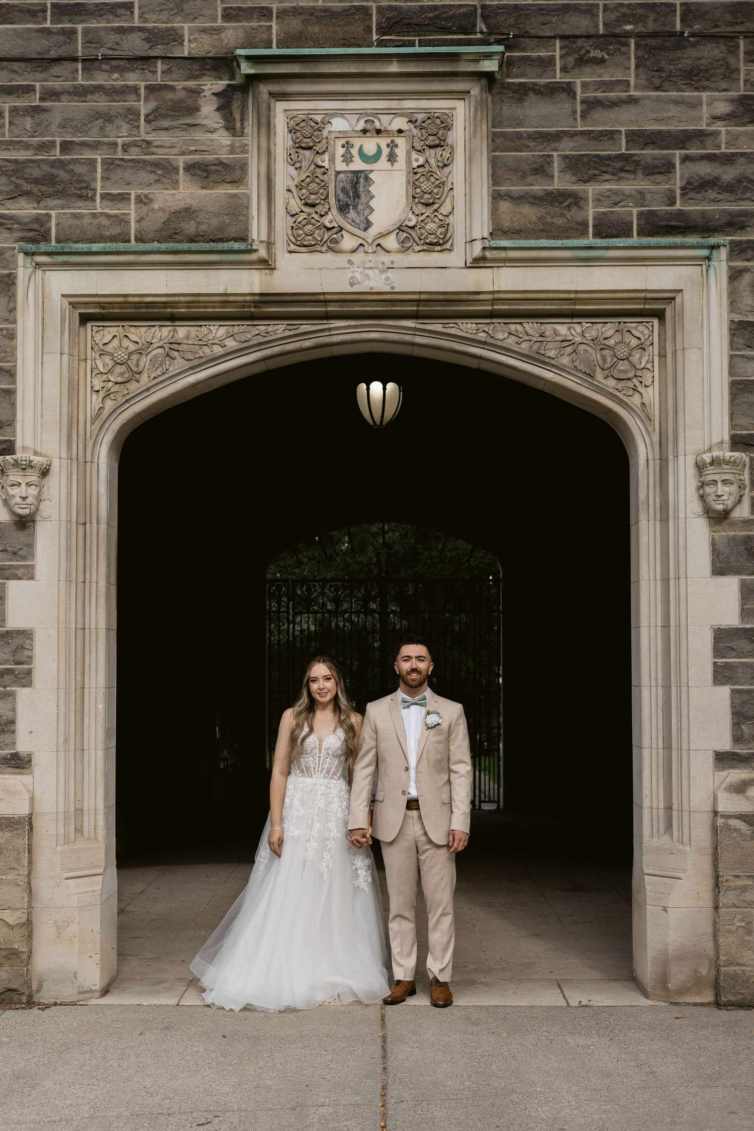 Wedding couple stands hand in hand beneath an ornate Gothic stone archway at a historic venue.