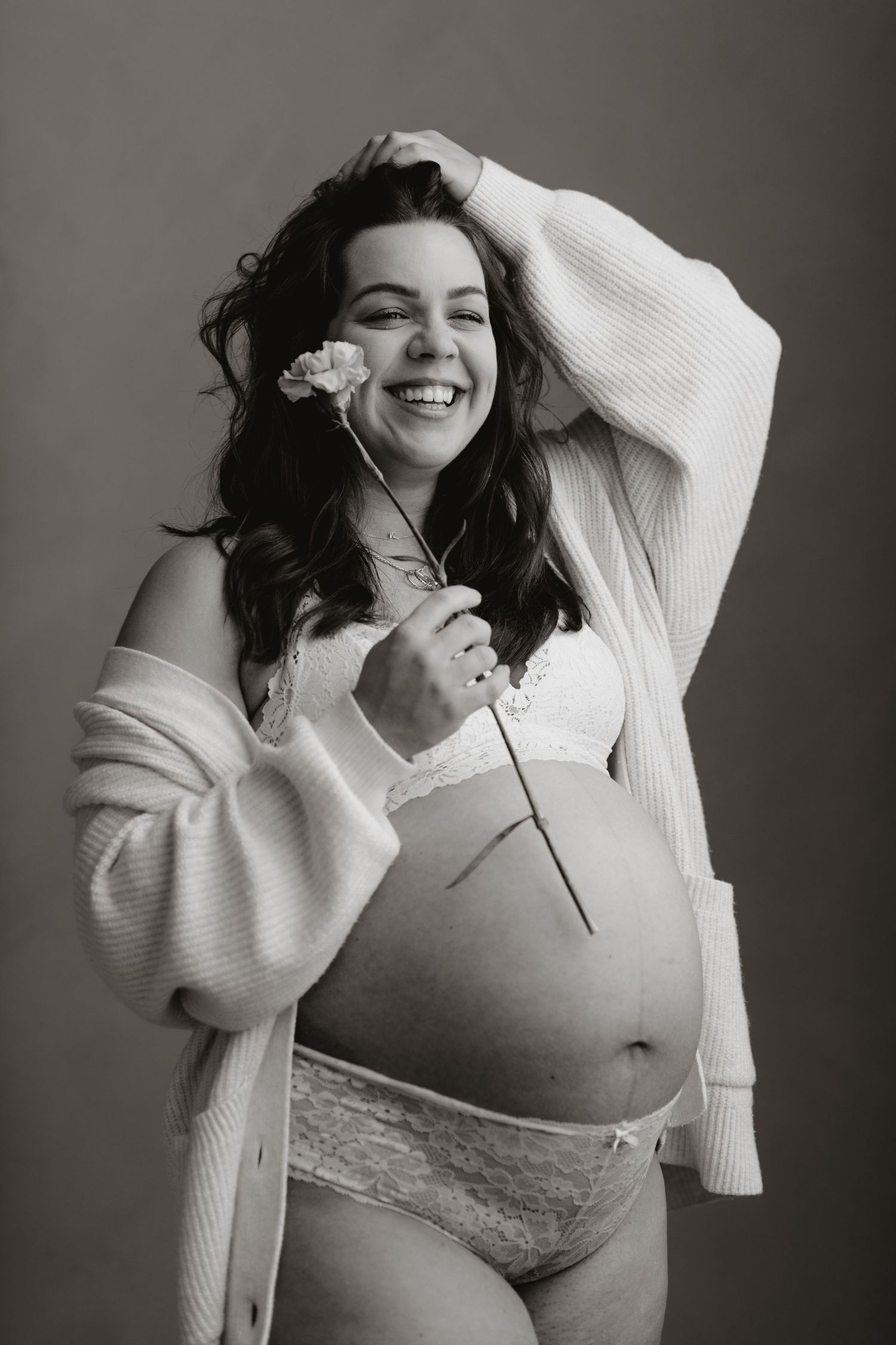 Black and white photo of laughing pregnant woman in lingerie holding flower during maternity session.