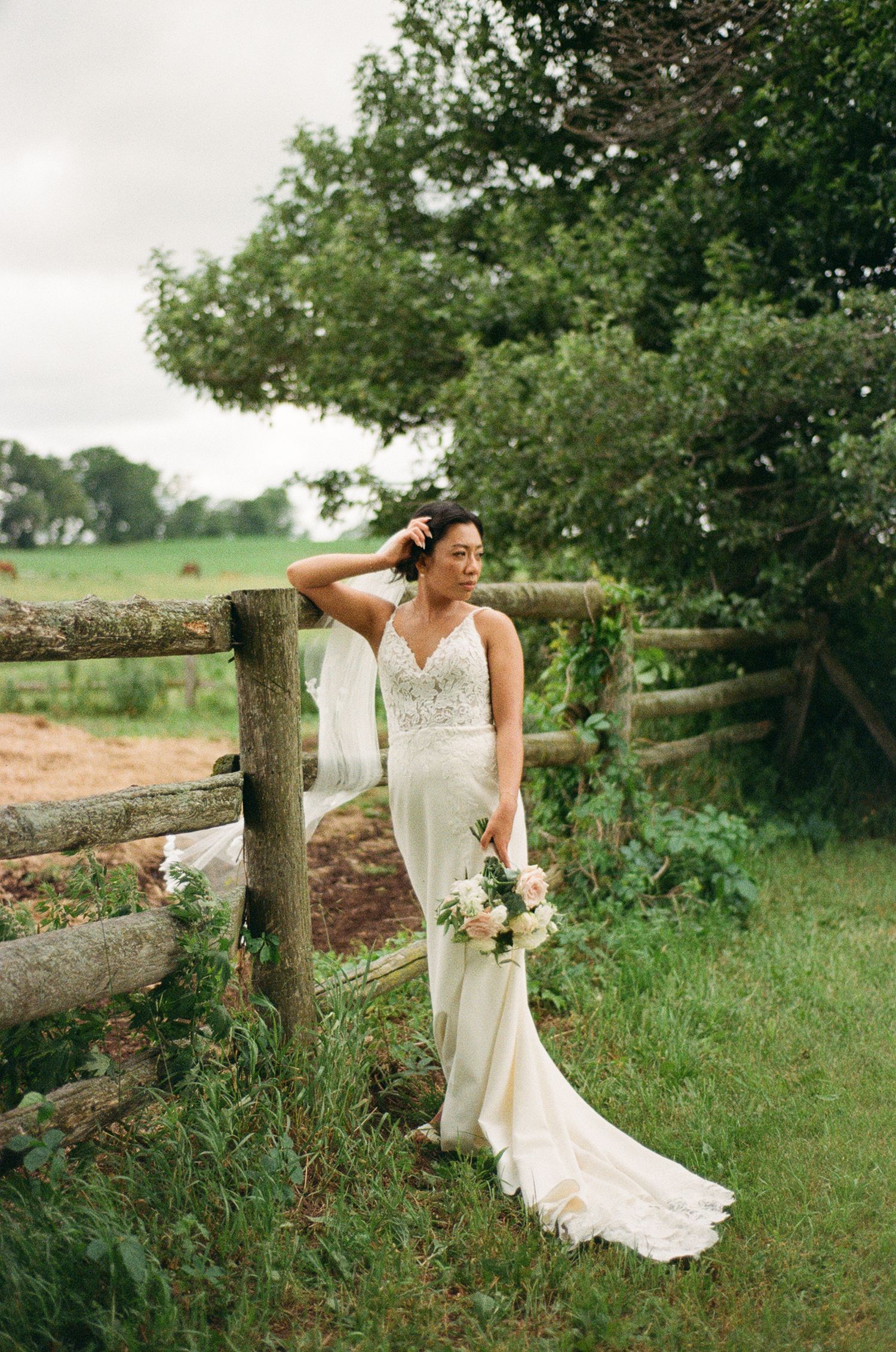 Bride in lace wedding gown holding bouquet leaning against rustic wooden fence surrounded by lush greenery on a farm.