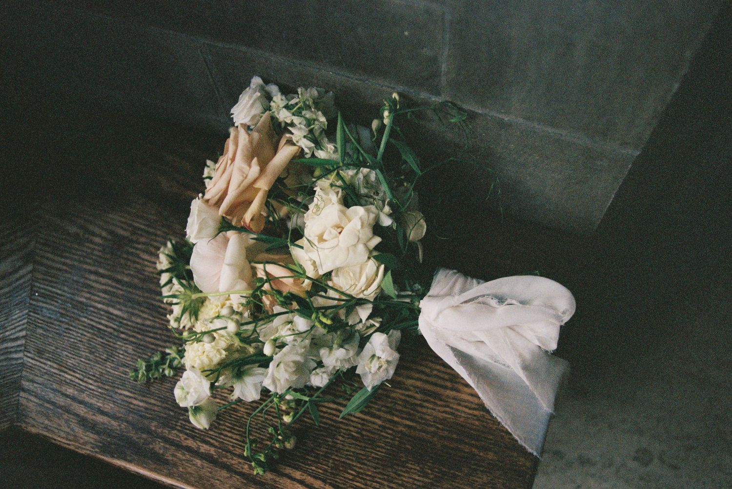White wedding bouquet with roses and greenery tied with silk ribbon resting on a wooden surface.