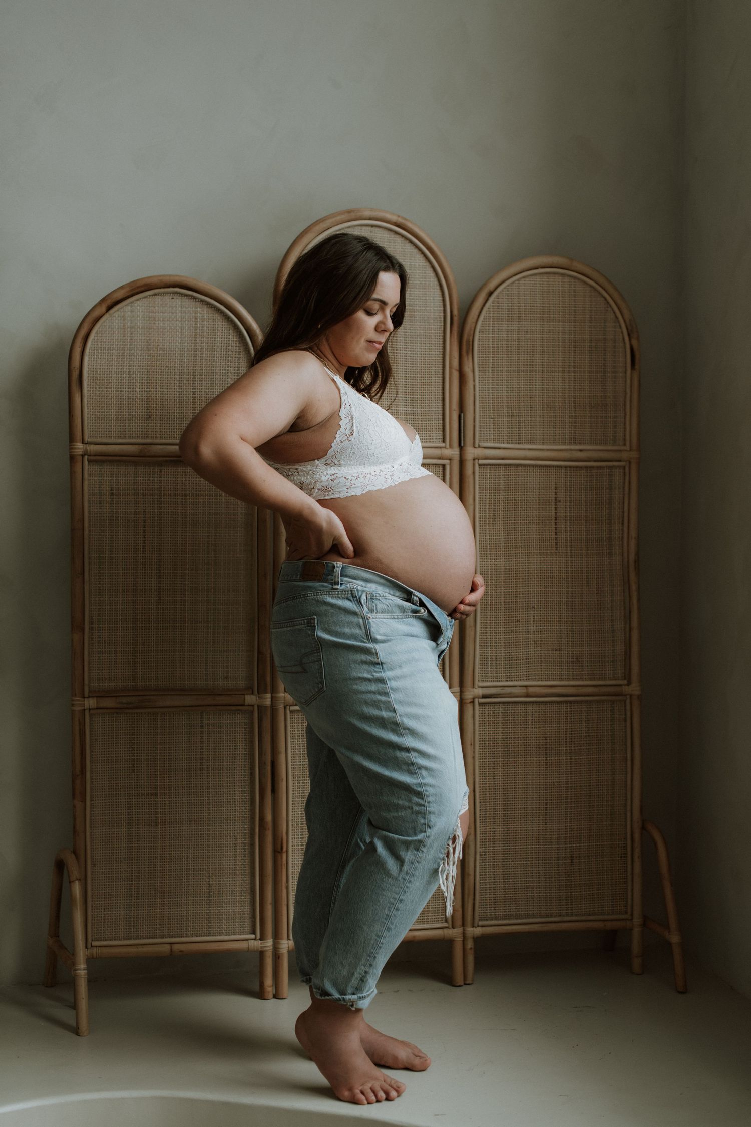 Pregnant woman in white lace bra and jeans leaning against a rattan room divider screen.
