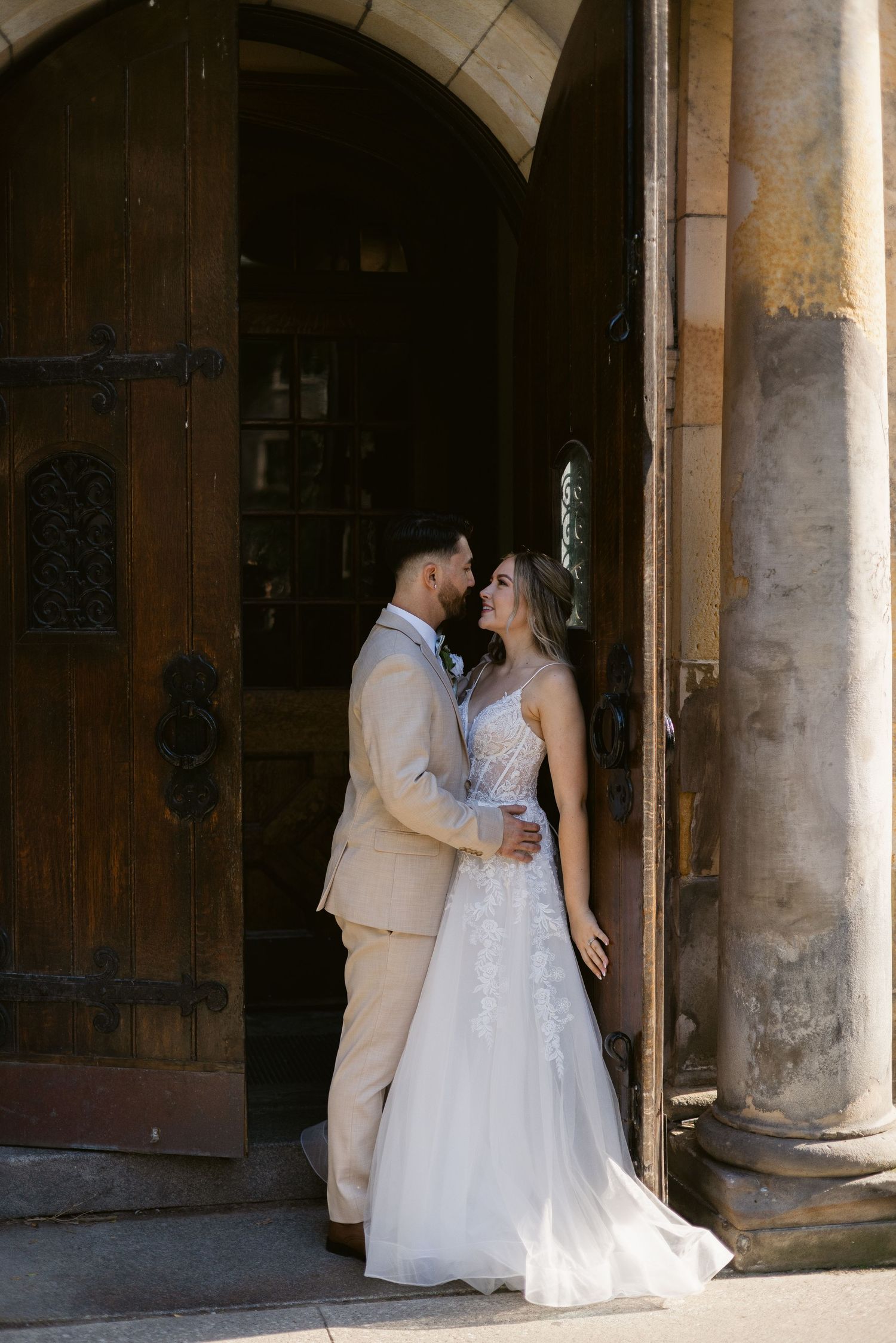 Bride and groom share romantic moment embracing in arched wooden doorway of historic stone building.
