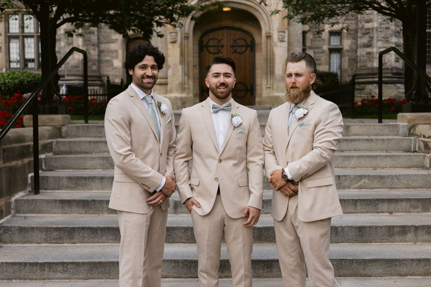 Three groomsmen in tan suits with boutonnieres pose casually on stone steps outside historic building.