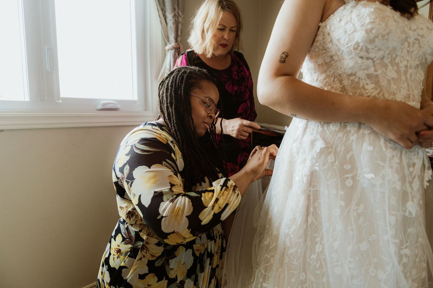 Two women helping bride into lace wedding dress, carefully fastening buttons before the ceremony.