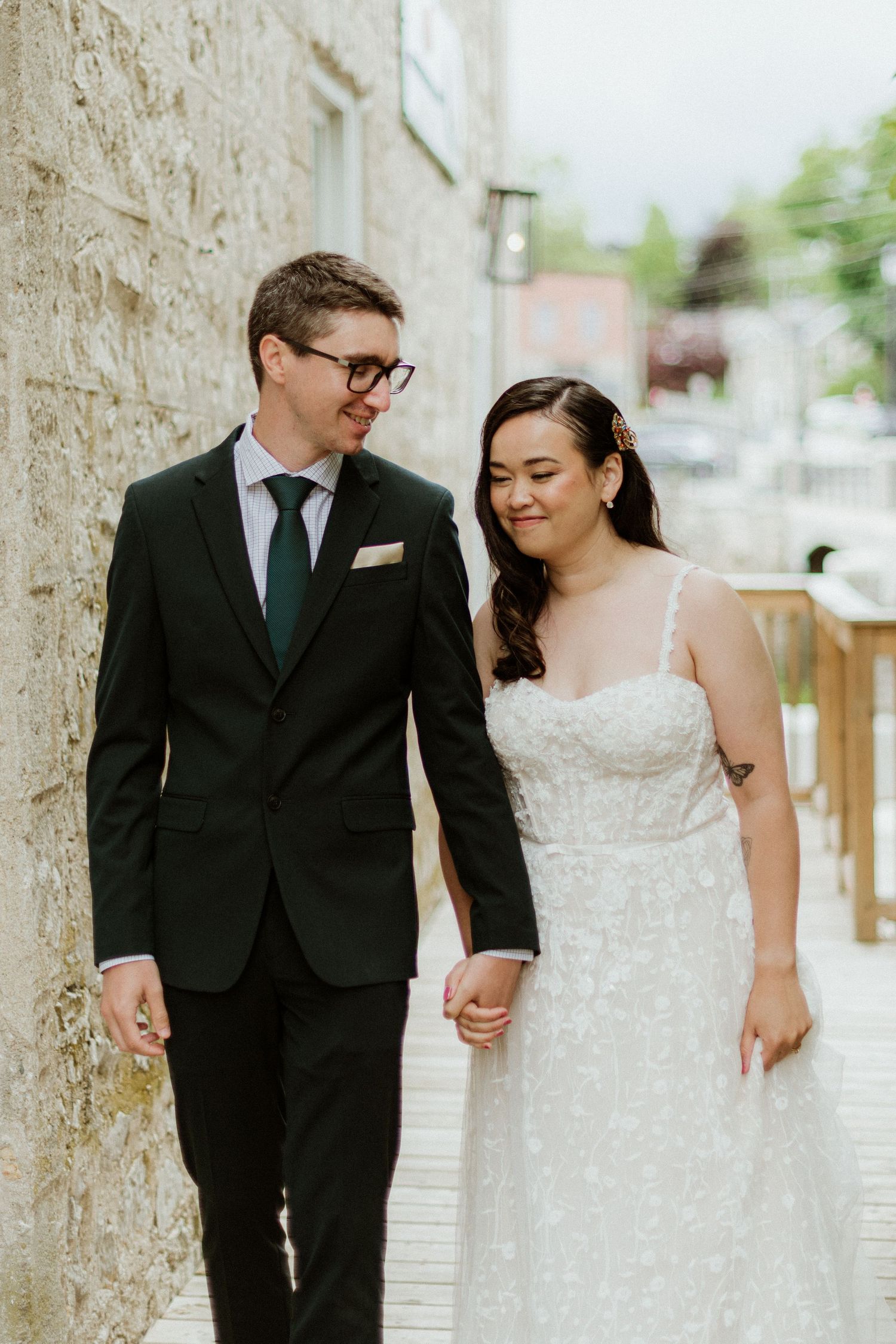 Bride in lace gown and groom in black suit holding hands while walking along a stone building.