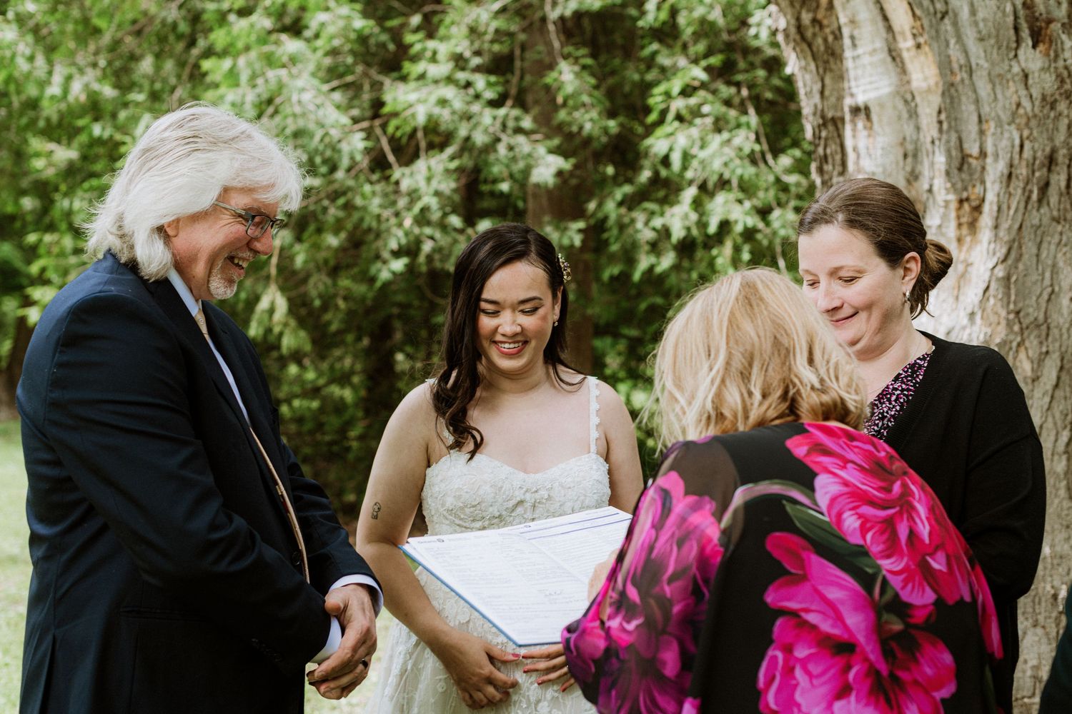 Bride smiling joyfully with wedding guests holding certificate near a large tree at outdoor ceremony.