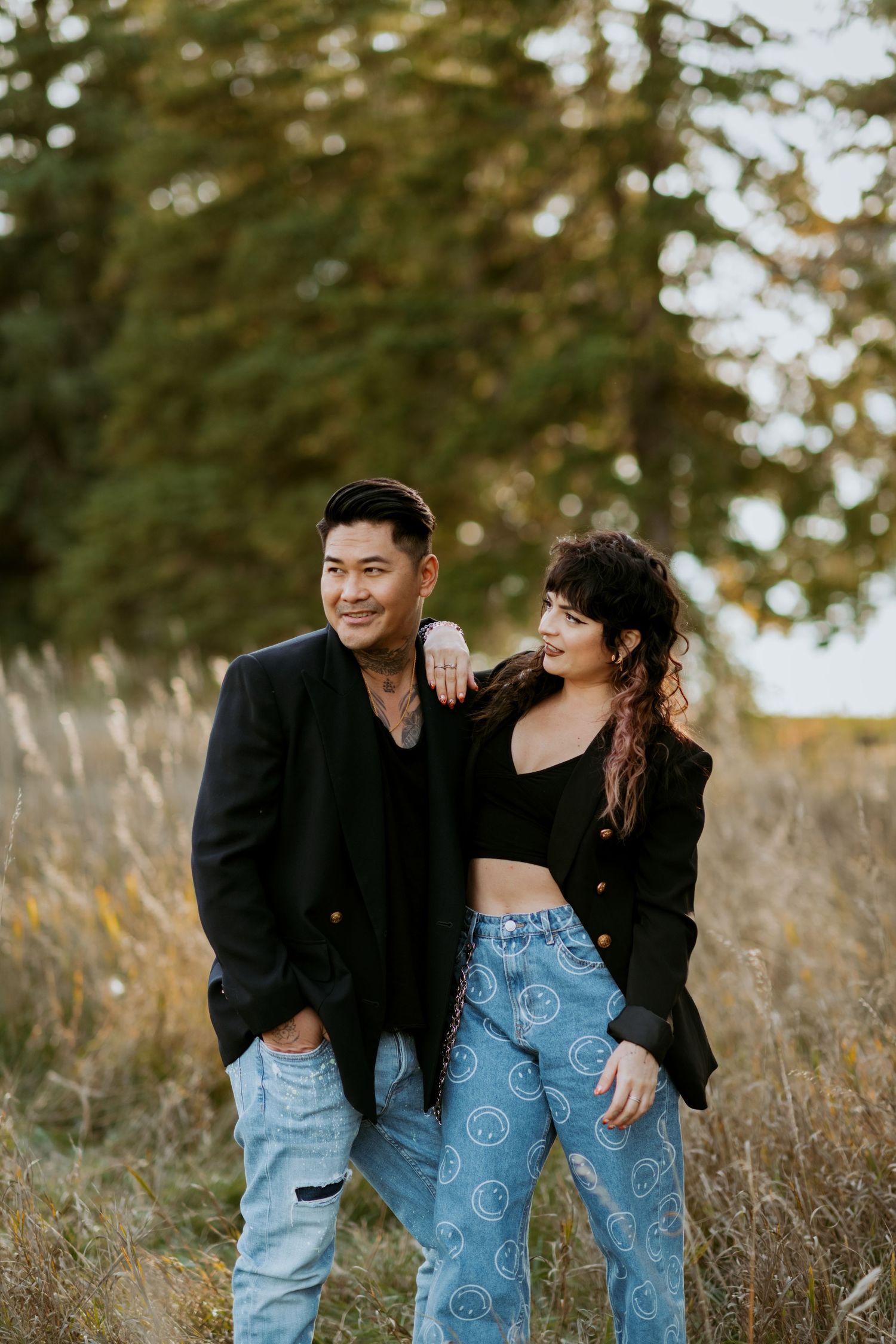 Smiling couple in black blazers and jeans standing together in a sunlit autumn field near pine trees.