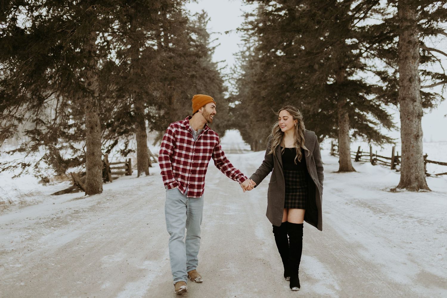 Happy couple holding hands walking down snowy tree-lined path in winter forest.