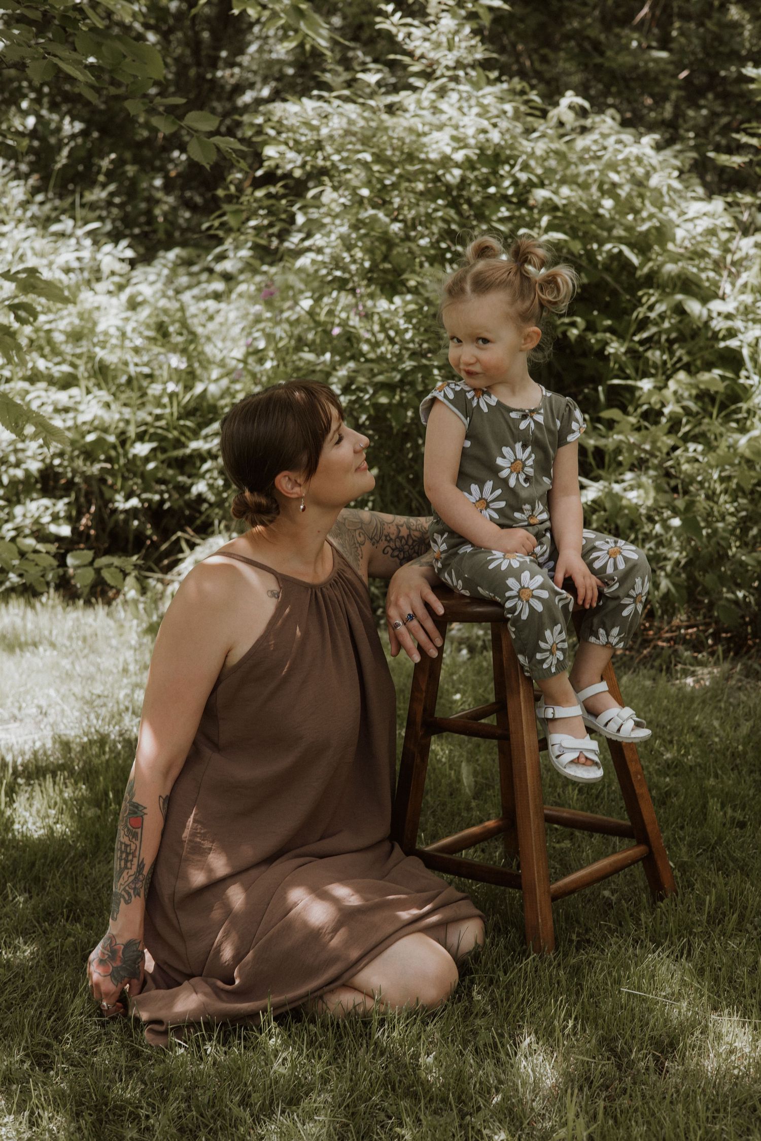 Tattooed mother in brown halter dress kneeling on grass with toddler on wooden stool outdoors.