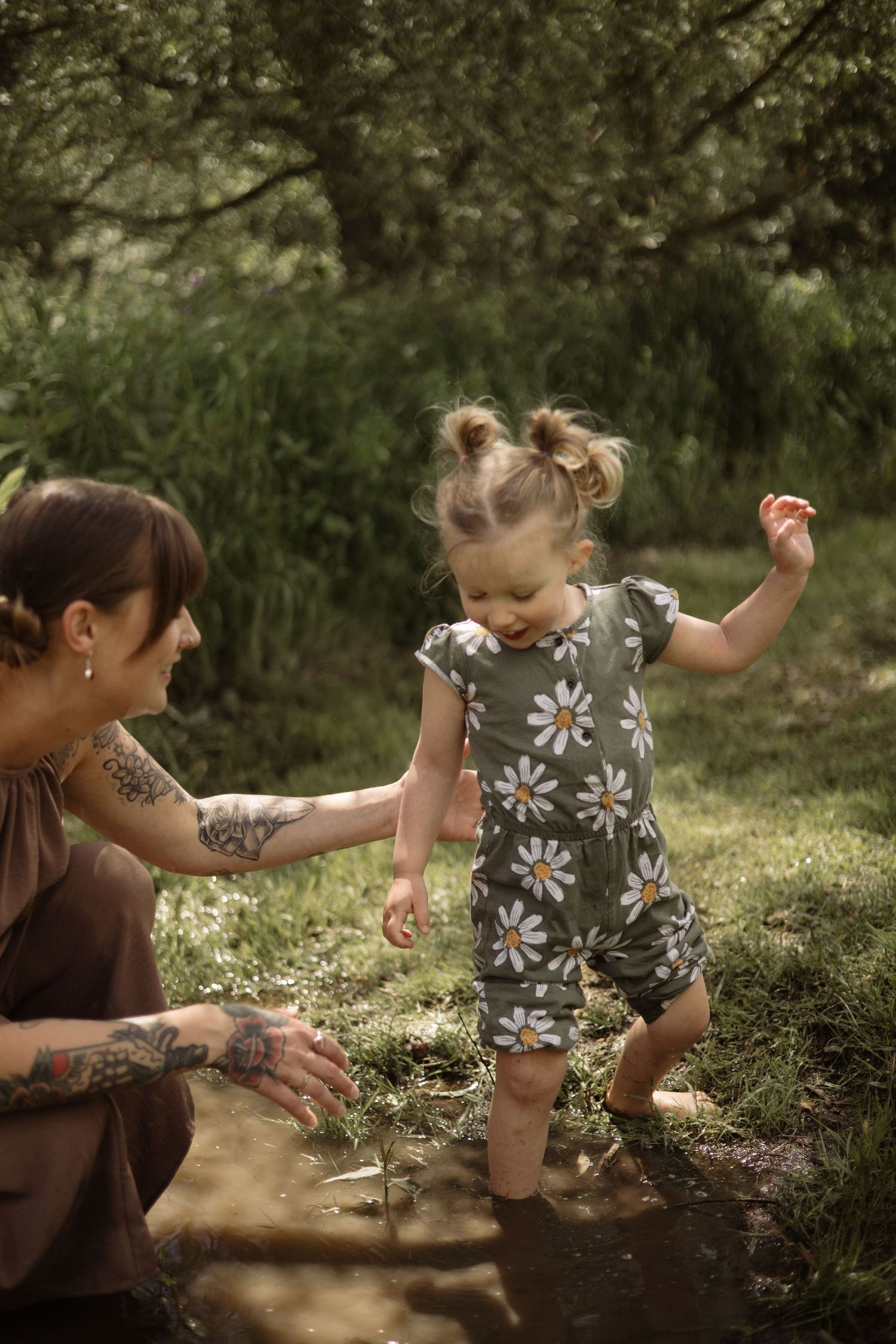 Tattooed mother smiling at her barefoot toddler daughter in a daisy romper splashing in a puddle outdoors.