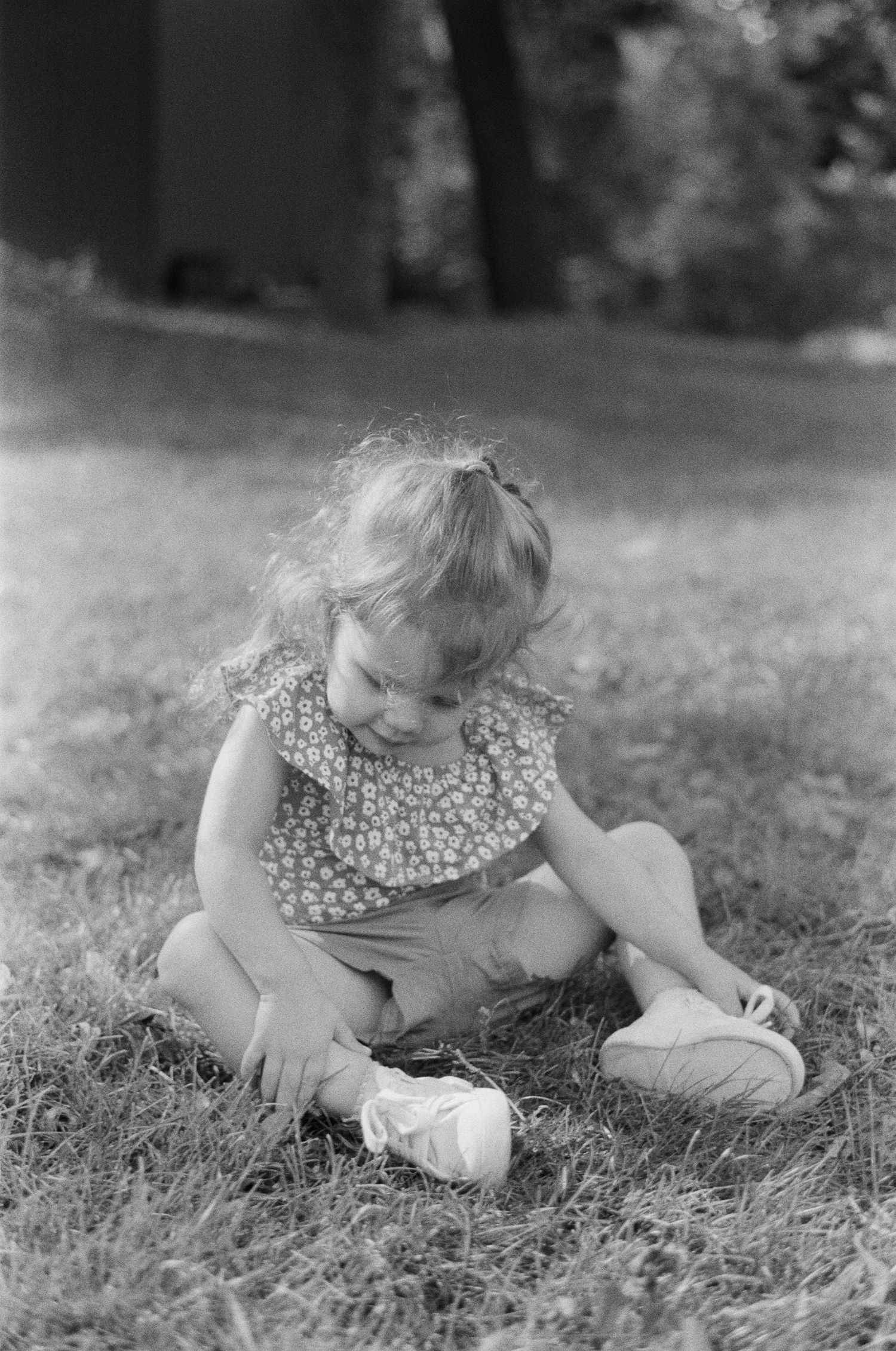 Black and white photo of a toddler in a ruffled floral top and shorts sitting on the grass and looking down at her white shoes.