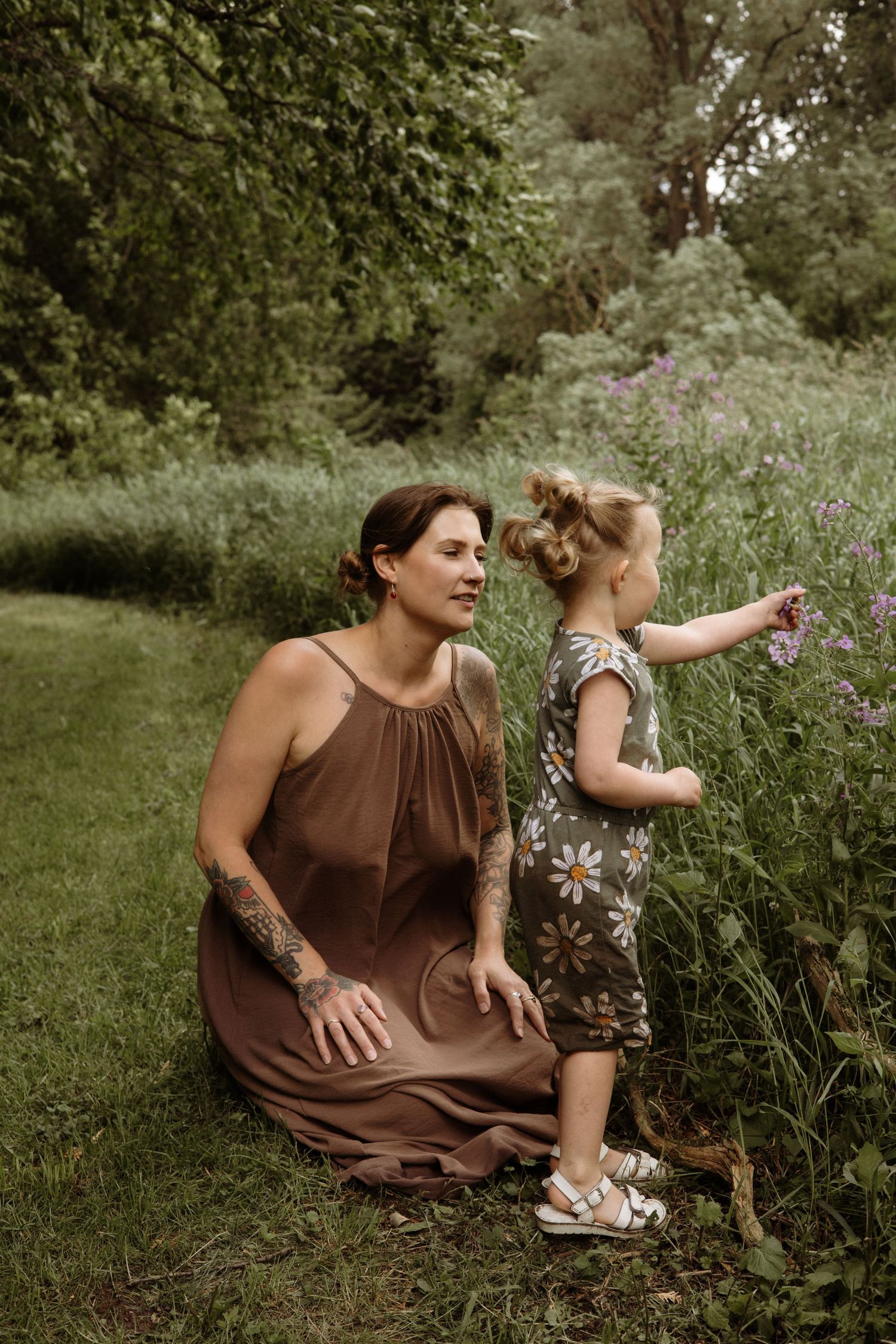 A tattooed mother in a brown dress sits on grass watching her young daughter pick purple wildflowers outdoors.