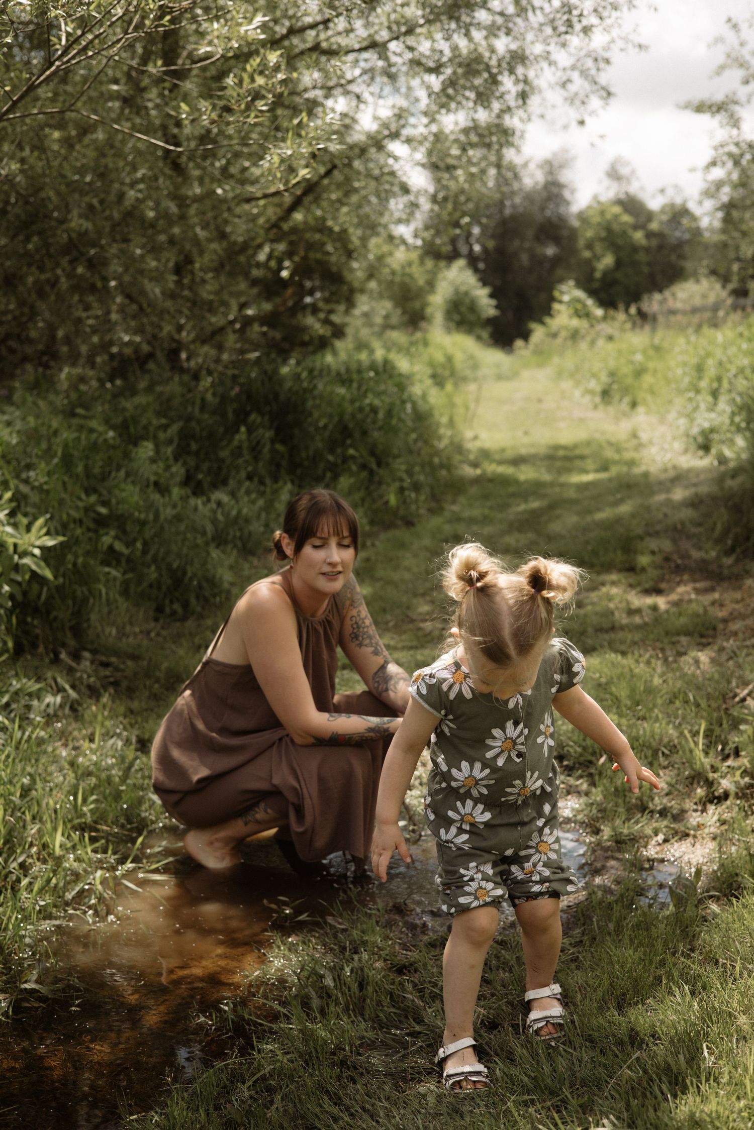 Mother in brown dress crouching in stream as toddler daughter in daisy print outfit splashes through the water outdoors.