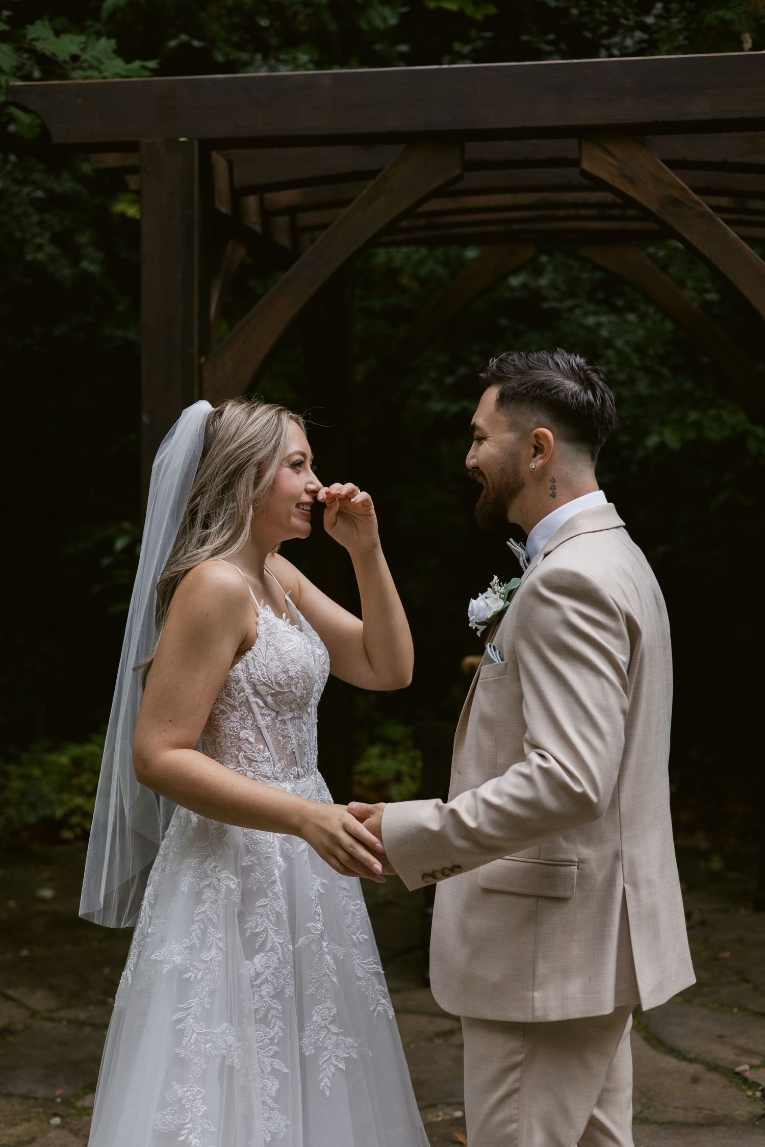 Bride wiping happy tears while holding hands with groom under wooden arch in garden setting.