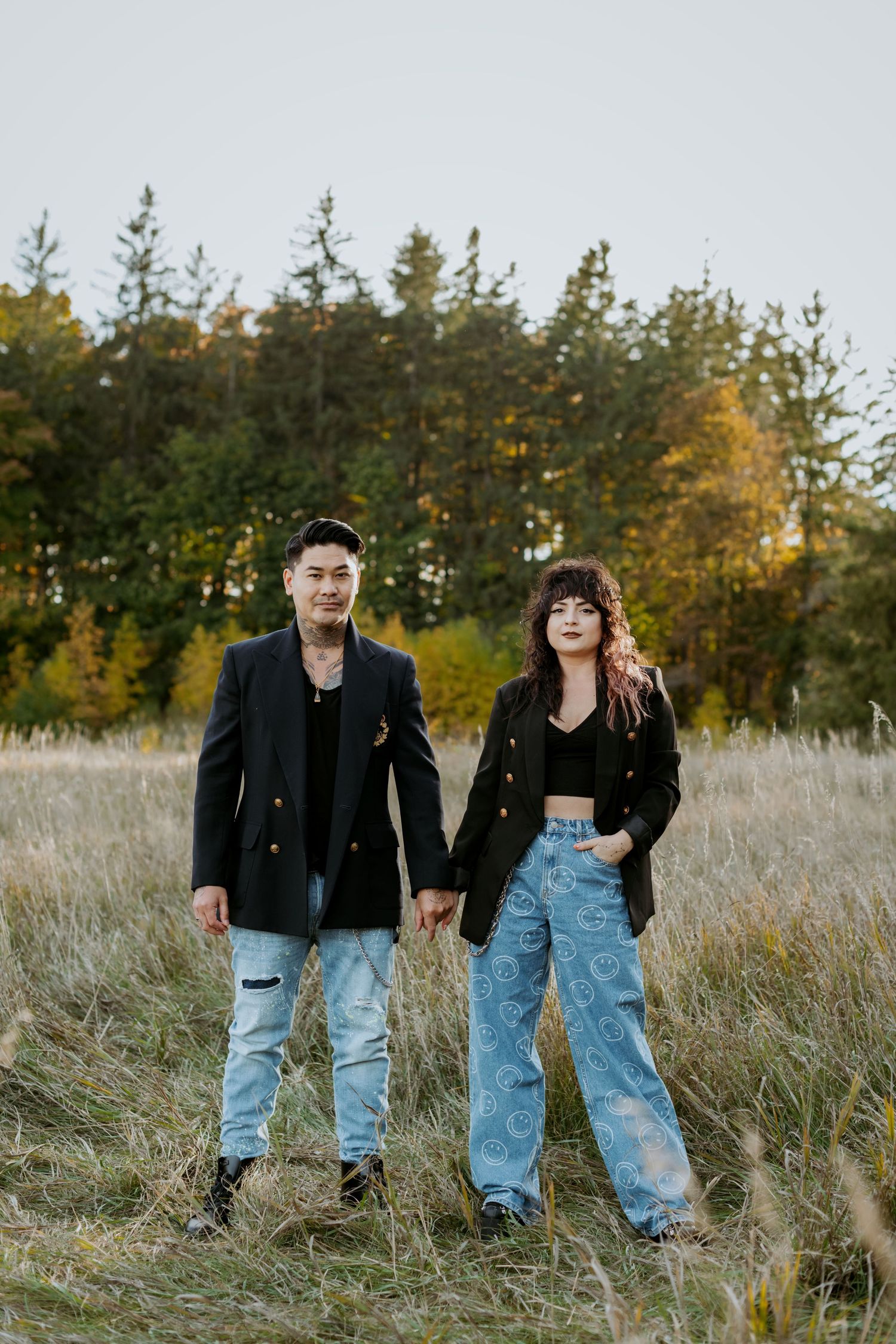 Stylish couple holding hands in autumn field wearing matching black blazers and ripped jeans with evergreen trees behind.