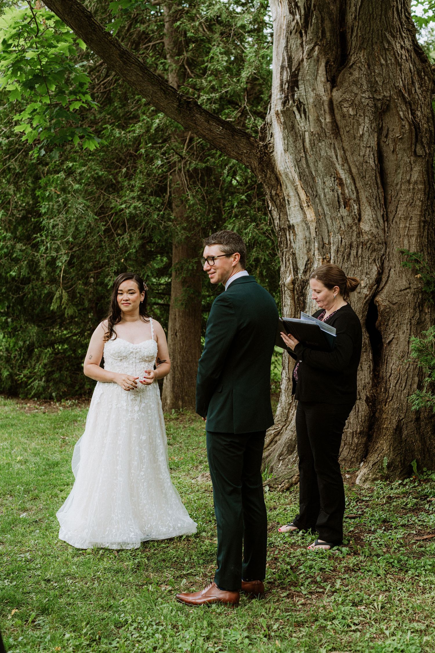Bride and groom exchange vows outdoors near a large tree with officiant during intimate wedding ceremony.